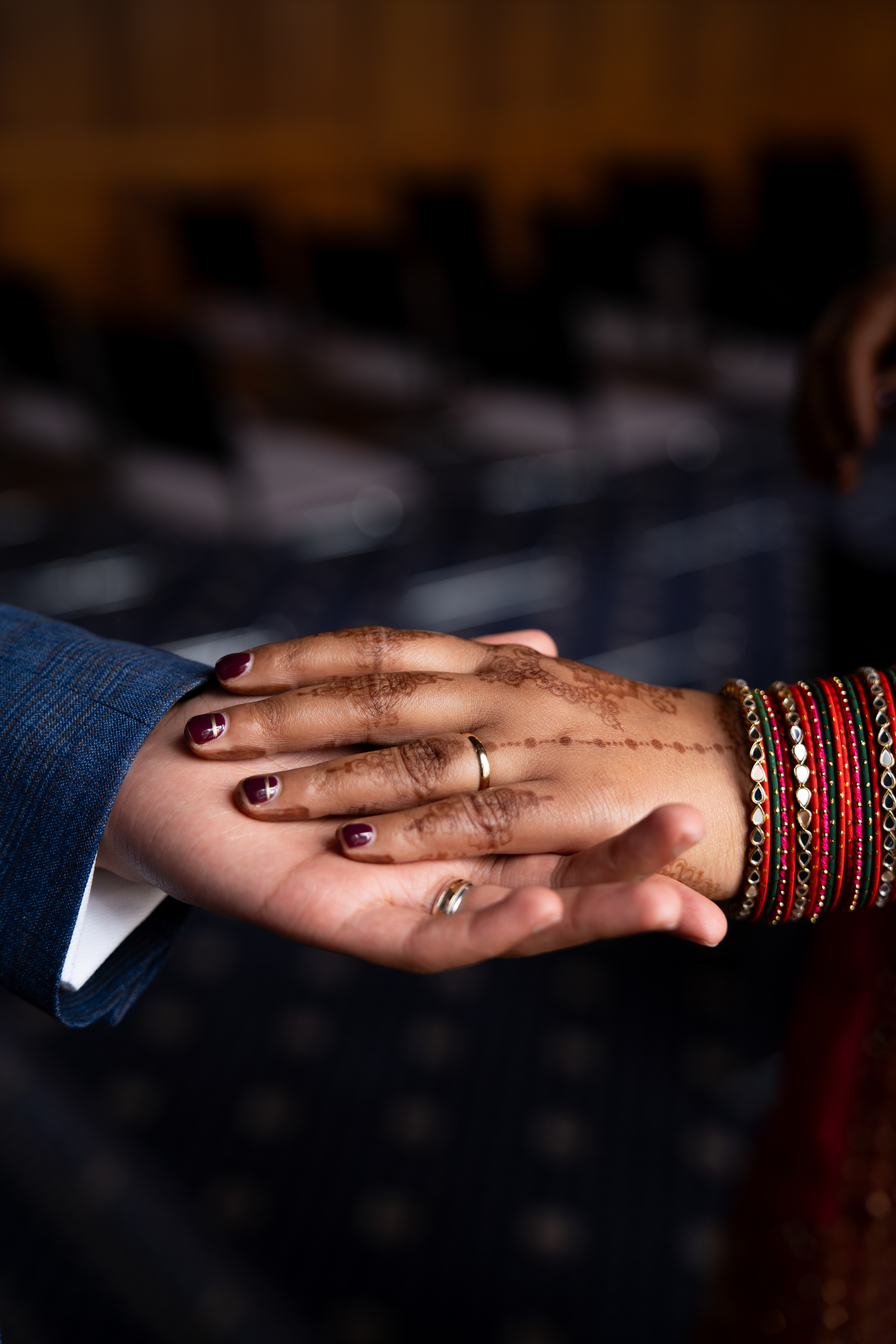 Two brown hands, both wearing wedding rings, are shown, loosely clasped. The woman's hand has mehndi and she wears a lot of colourful bracelets.