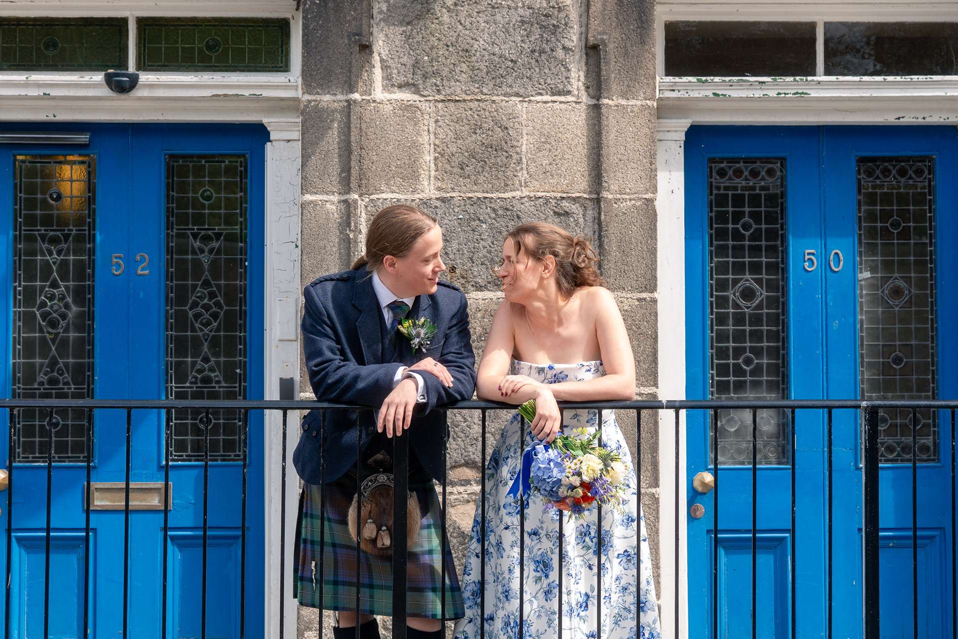 A long-haired man in a blue kilt and a woman in a blue floral wedding dress, holding a colourful bouquet, lean over a railing, arms loosely draped, smiling at each other. Behind them are two blue wooden doors with stained glass panelling.