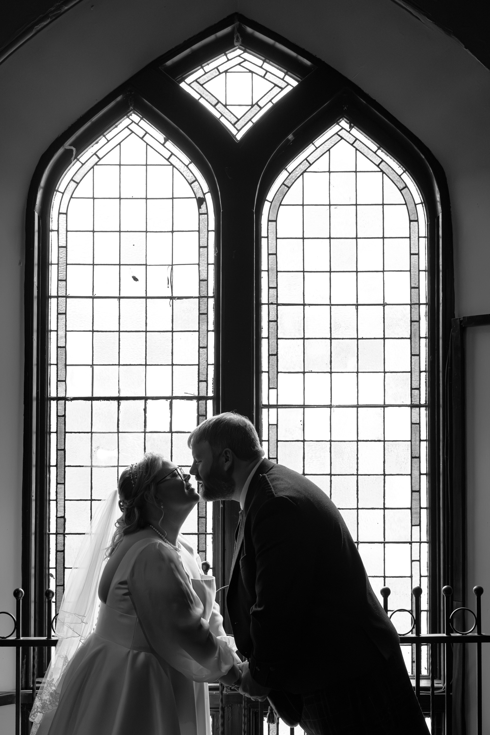 Slains Aberdeen wedding: Black and white photo of bride and groom leaning in for a kiss in front of stained glass window.