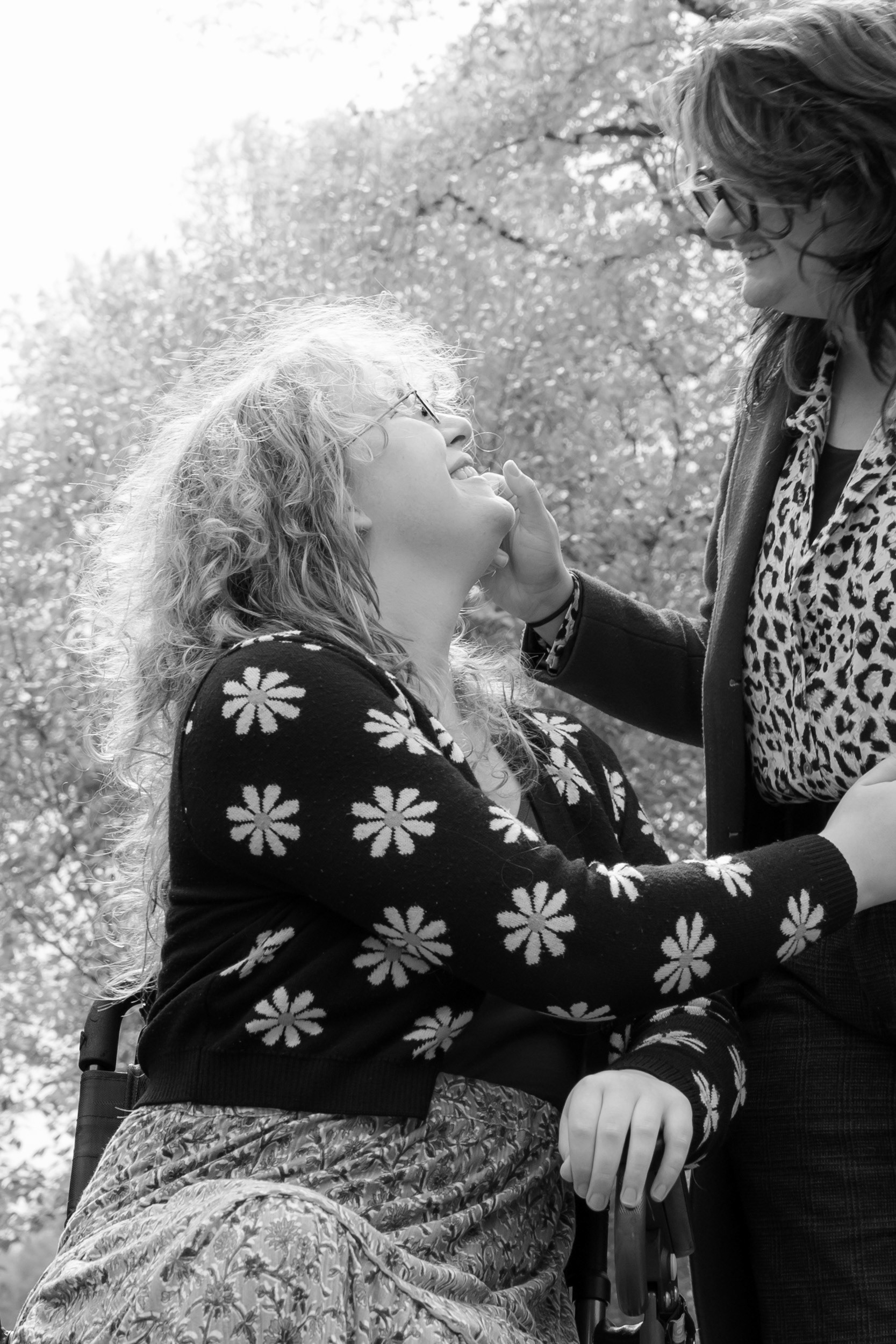 Young woman in floral cardigan sits in her wheelchair, glowing as she is backlit by the sun. Her partner reaches down to touch her face and they are beaming at each other.