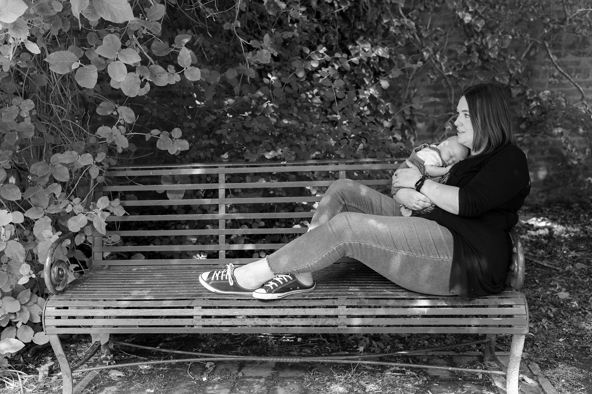 Aberdeen Newborn Photography: A black and white image of a mum stretched out on a park bench, shaded by creepers, as her baby sleeps snuggled on her chest.