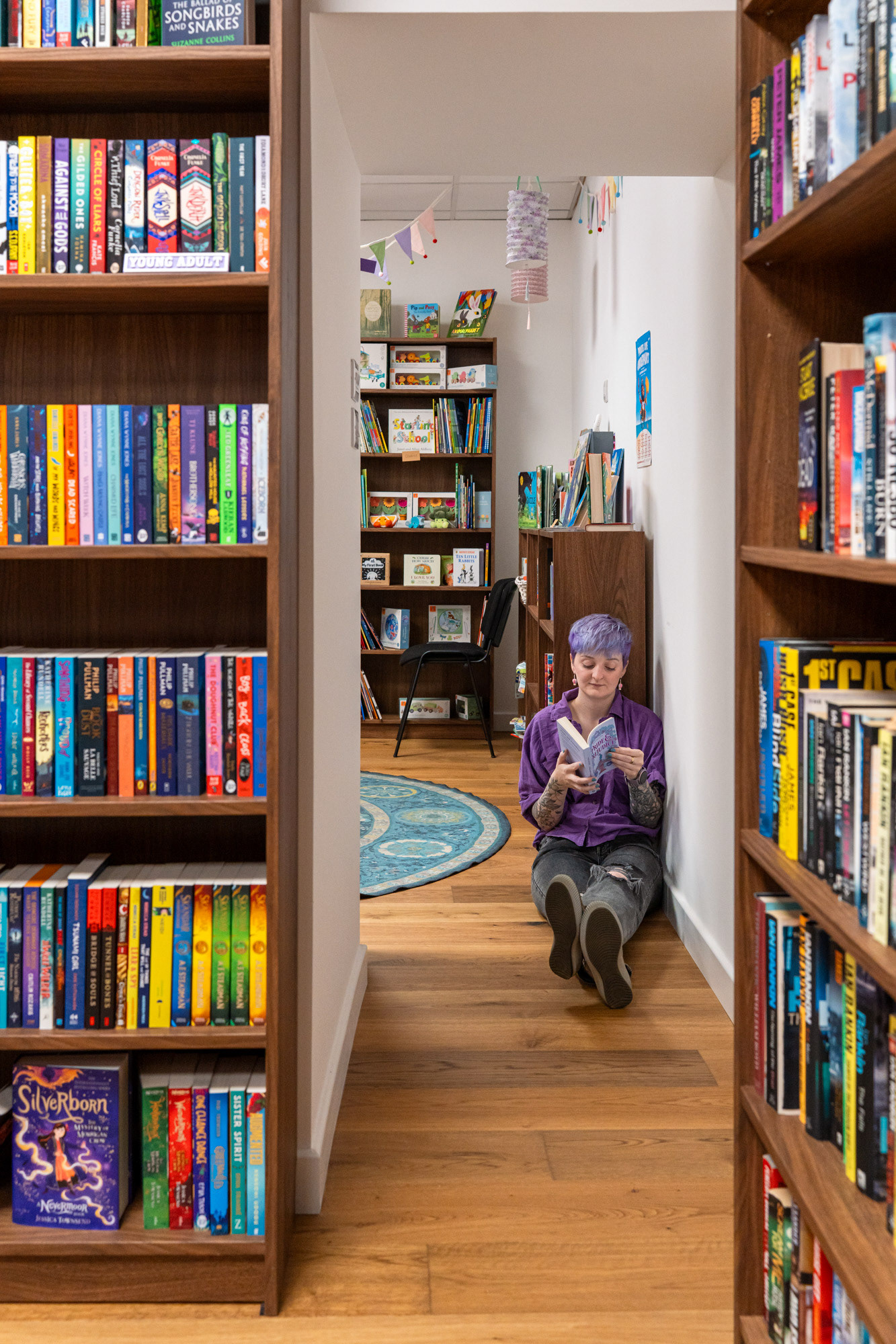 Aberdeen branding photography for Somerville Books: Owner, Christie, dressed in purple shirt and jeans, with purple hair, is seen through a gap between shelves, sitting on the floor, reading a purple book.
