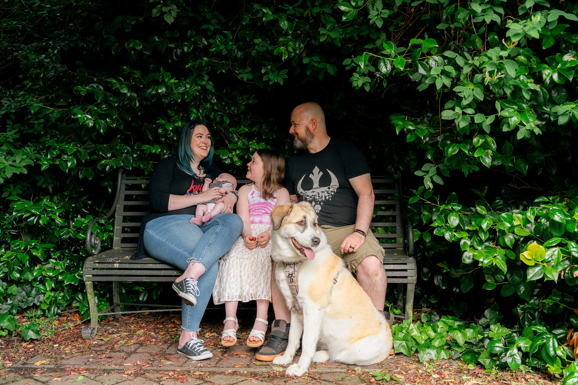 A family of four beams at each other during their alternative newborn photo shoot. They are gathered on a metal park bench, completely surrounded by shrubs and creepers. Their large dog sits in front of them.