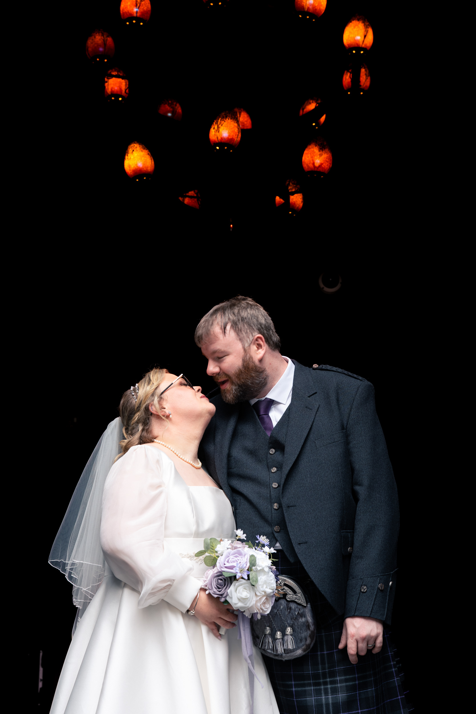 Alternative Aberdeen wedding photography: Bride and groom lean in for a kiss with a glowing orange chandelier above them.