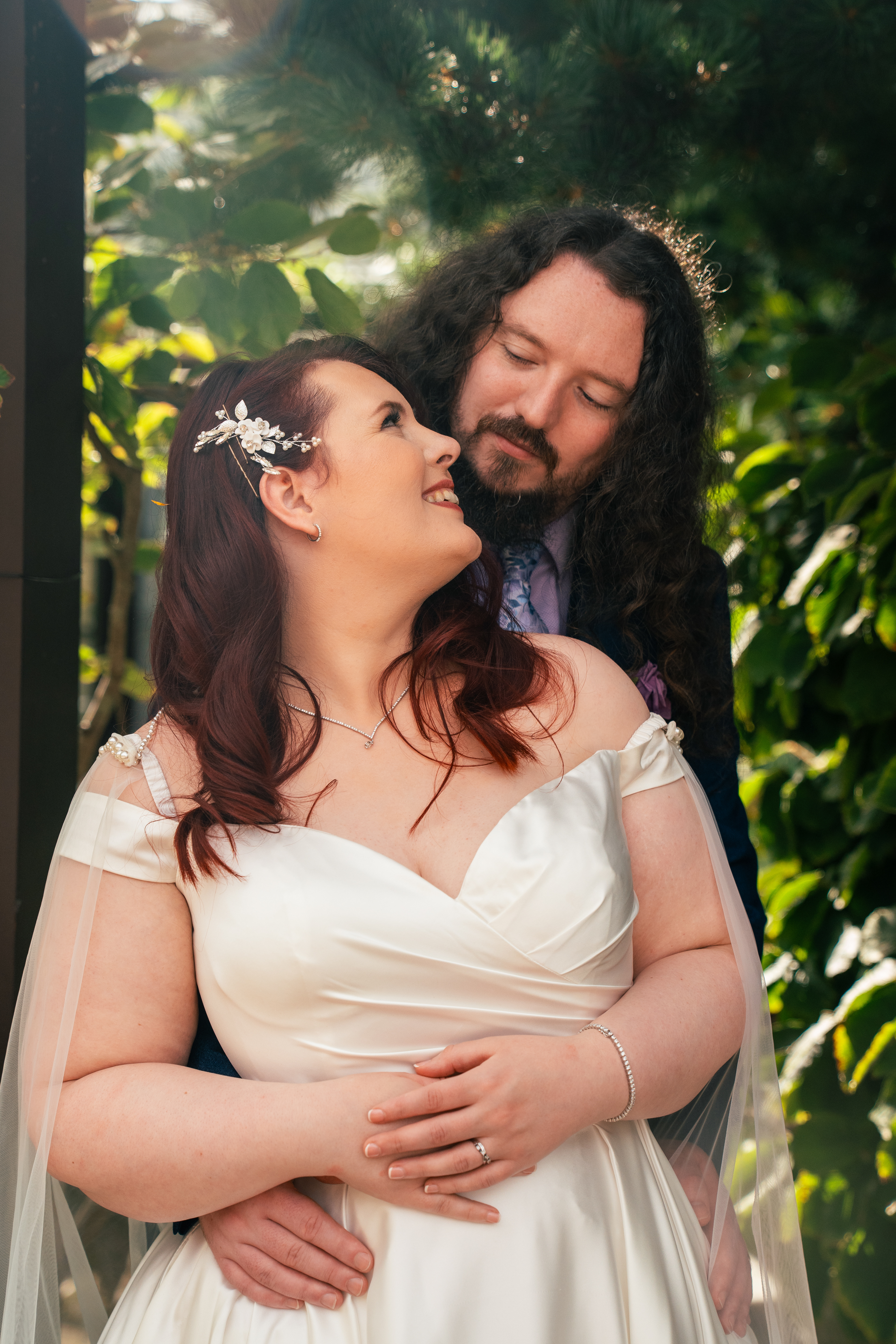 A dark haired bride in cream dress looks back at her long-haired husband as he hugs her from behind. The background is greenery, with sun flare dappling the scene.