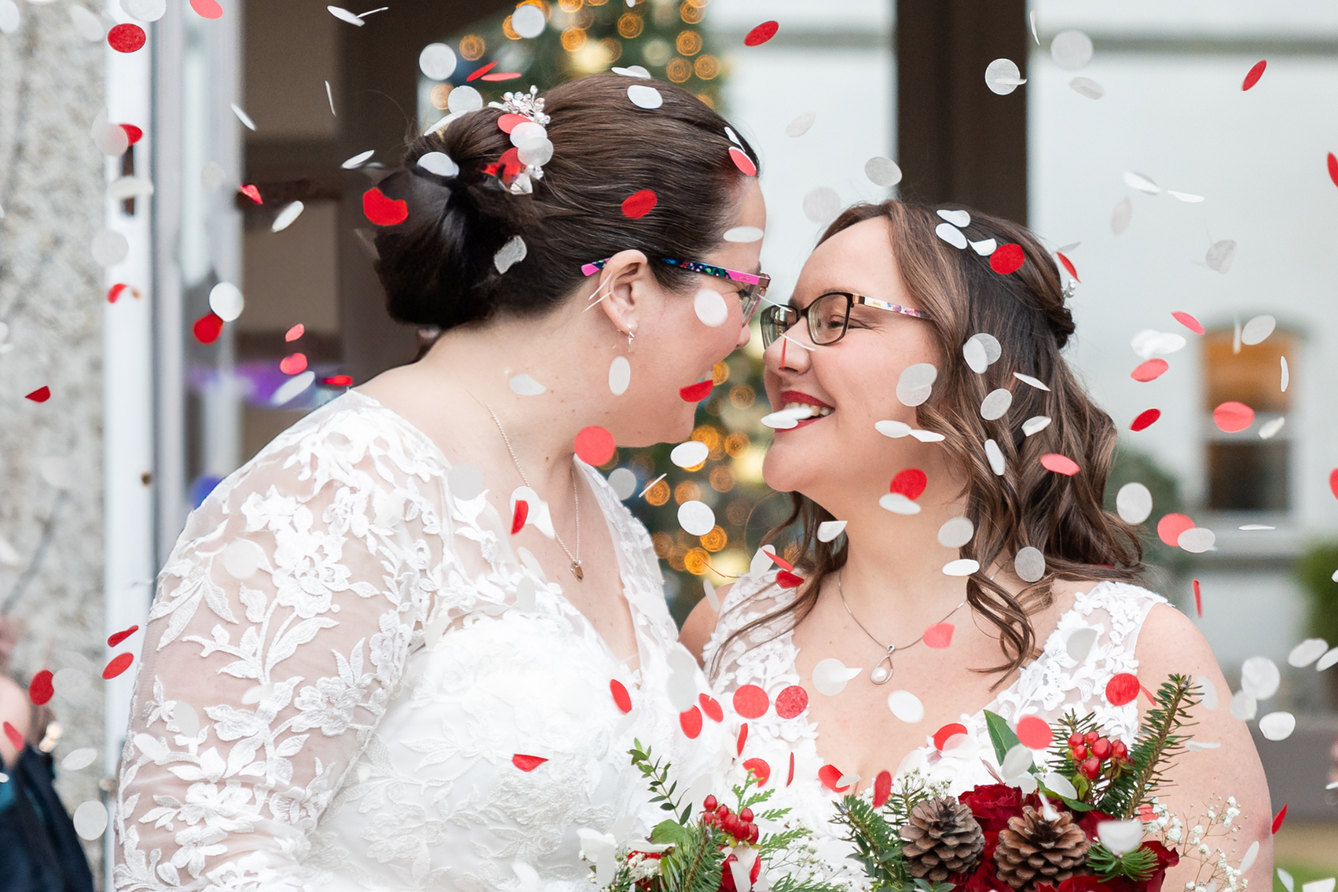 Two brides, both brunette and white skinned, wearing white wedding dresses and carrying bouquets of red roses, lean in for a kiss while red and white confetti drifts around them.