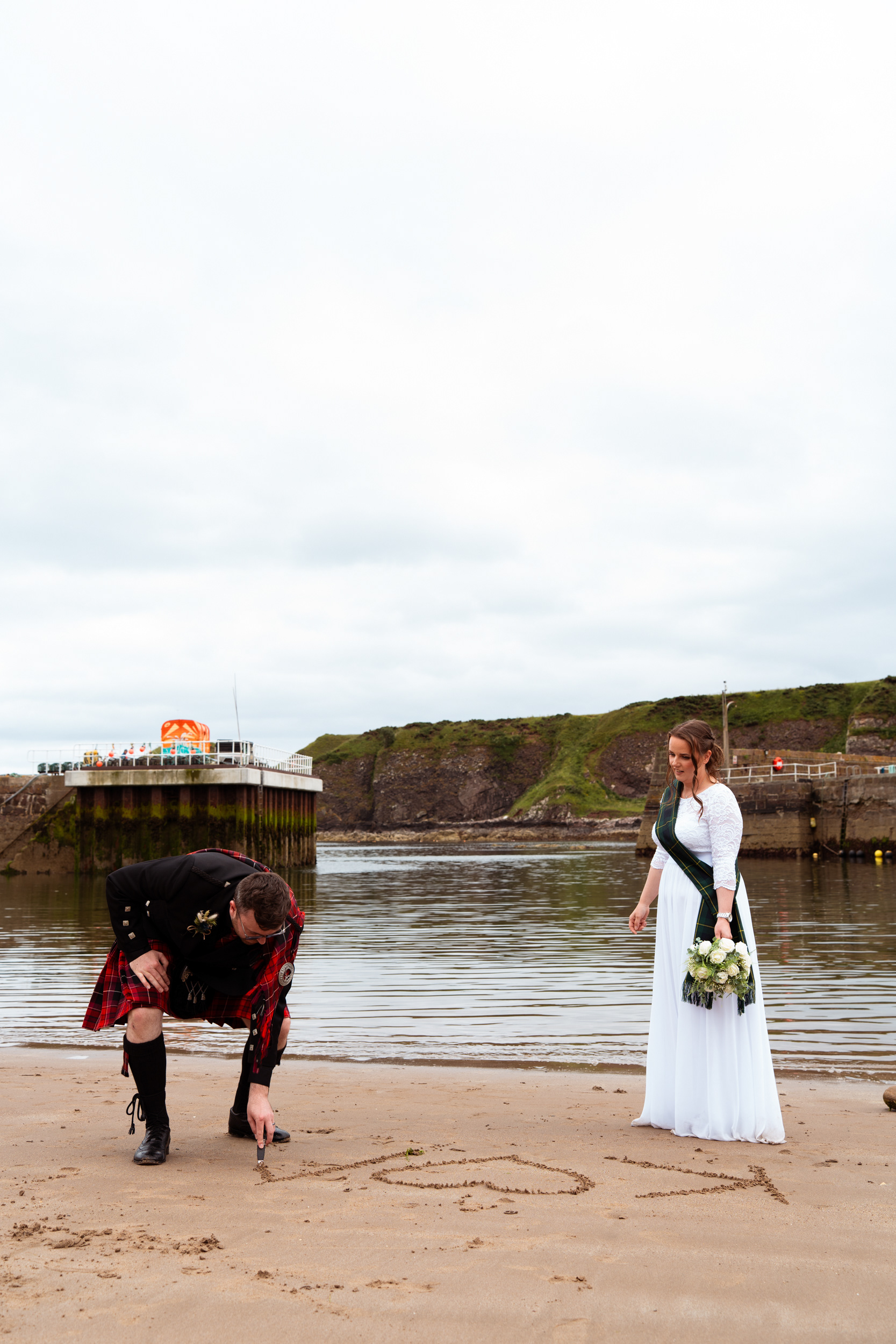 Alternative Aberdeenshire wedding photography: Groom in red kilt uses his sgian dubh to write initials in the sand at Stonehaven as his new wife looks on.