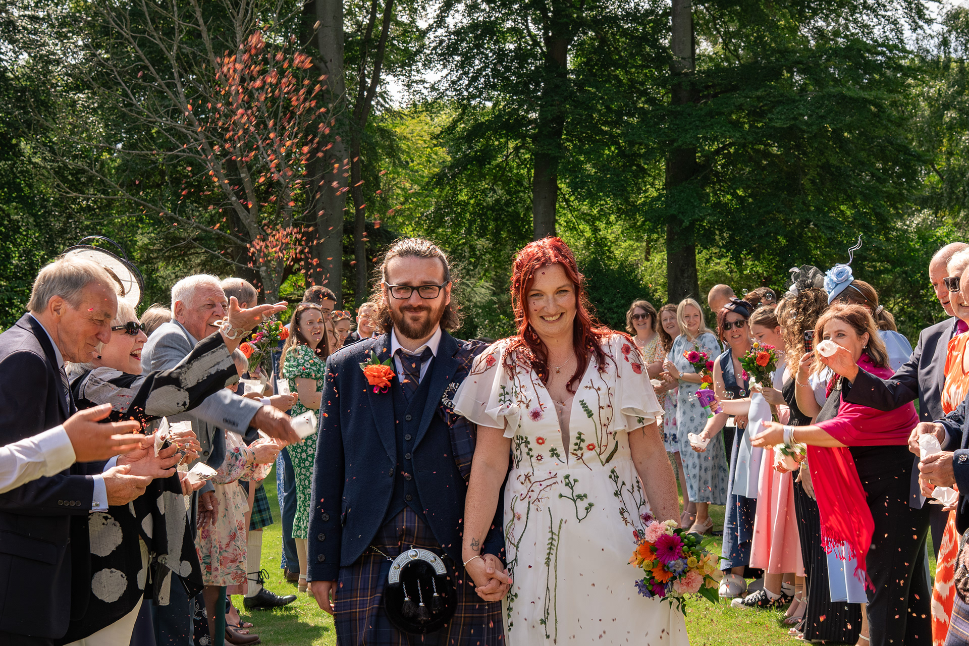 Bride and groom laugh as they are showered with red confetti, walking between their guests on a sunny day in Hazlehead Park