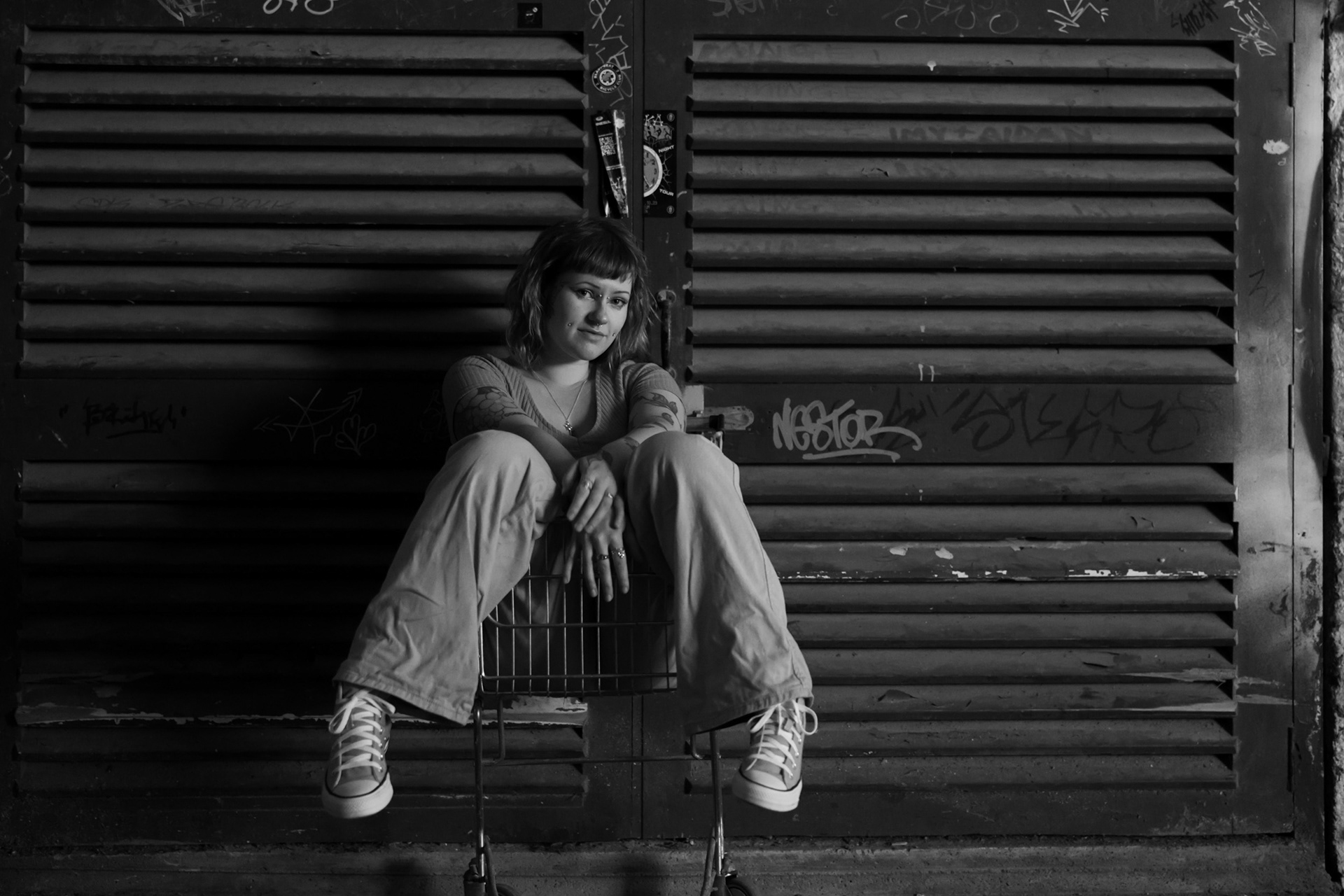 A black and white photo of a young woman sitting in a trolley, looking directly at the camera. The backdrop is of a dusty, slatted warehouse door.