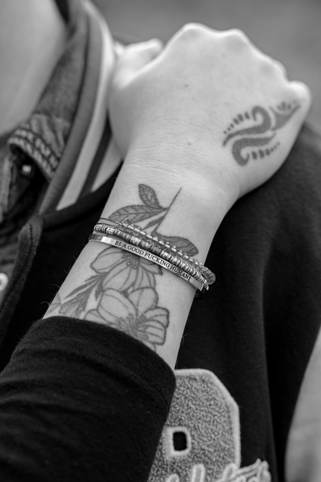 Close up of a tattooed wrist with a bracelet engraved with "BE A GOOD FUCKING HUMAN"
