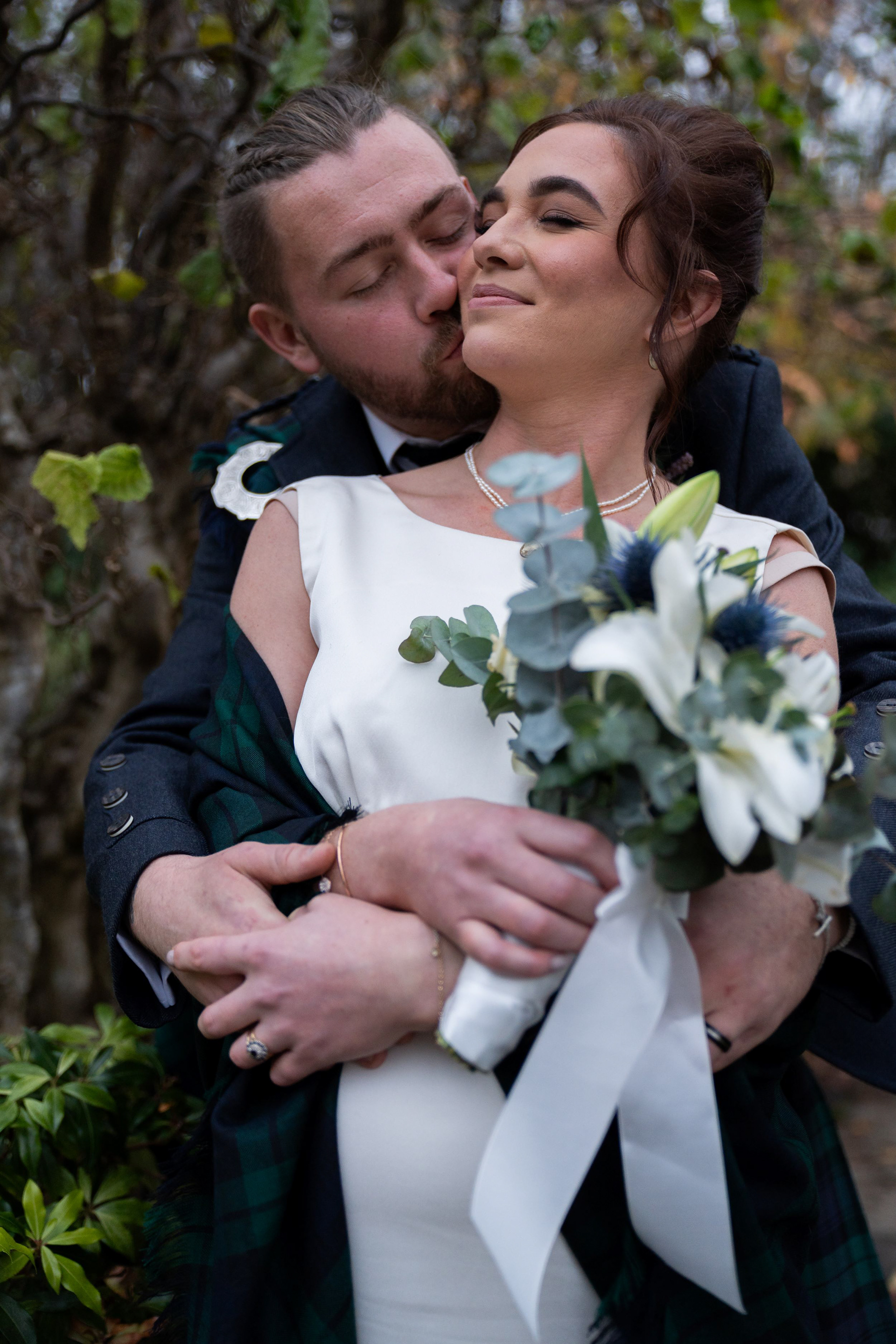Aberdeen Wedding Photography at Duthie Park Winter Gardens: A beautiful bride in white with tartan shawl wrapped around her closes her eyes as her groom in kilt with plaited hair style hugs her from behind and kisses her cheek.