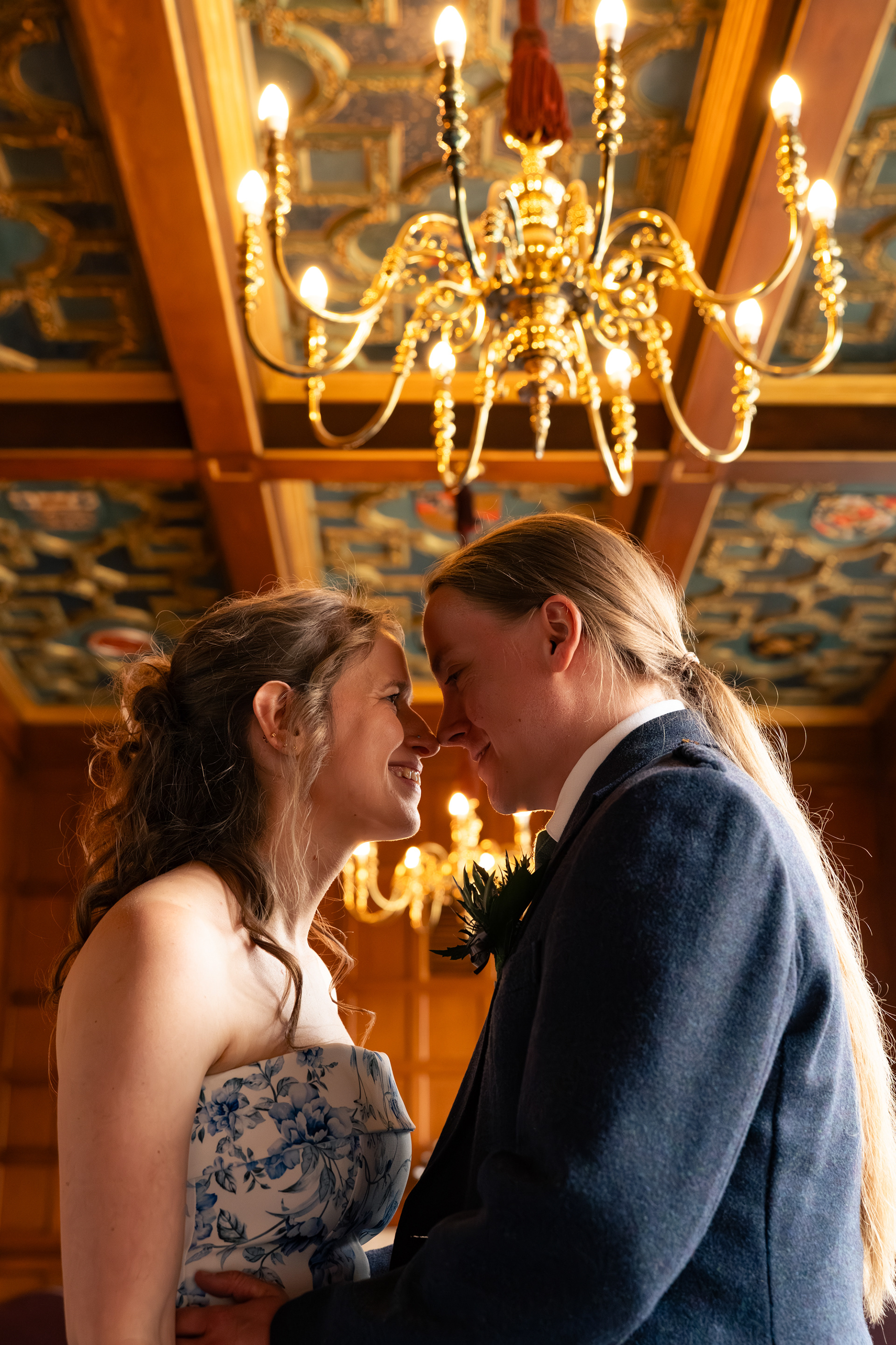 A bride in floral dress and long-haired groom in kilt stand face to face under the chandeliers and ornate ceiling of the Grant Room.