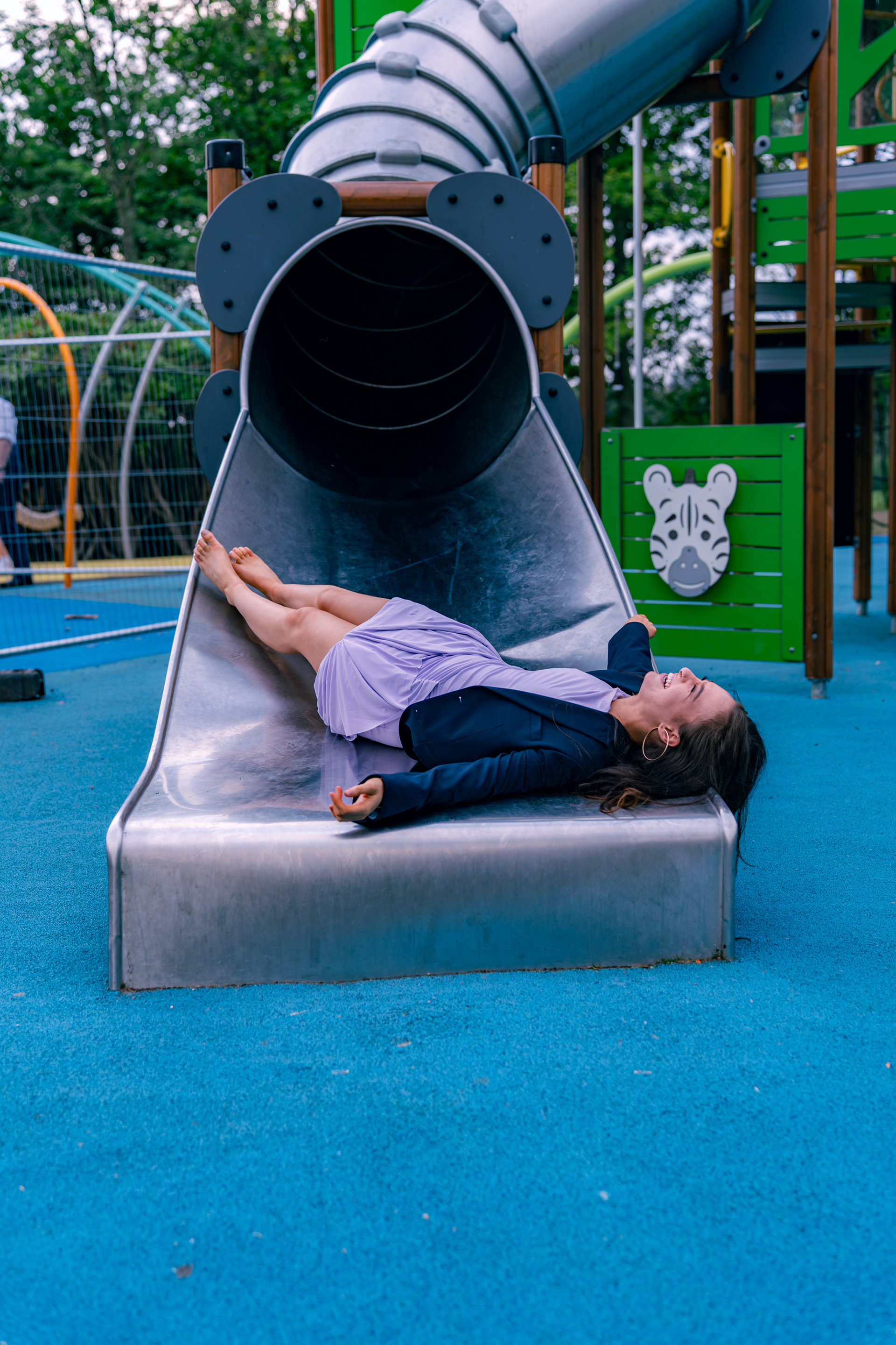 A young woman in a lilac dress and navy jacket lies, laughing, at the bottom of a tube slide during an Aberdeen wedding