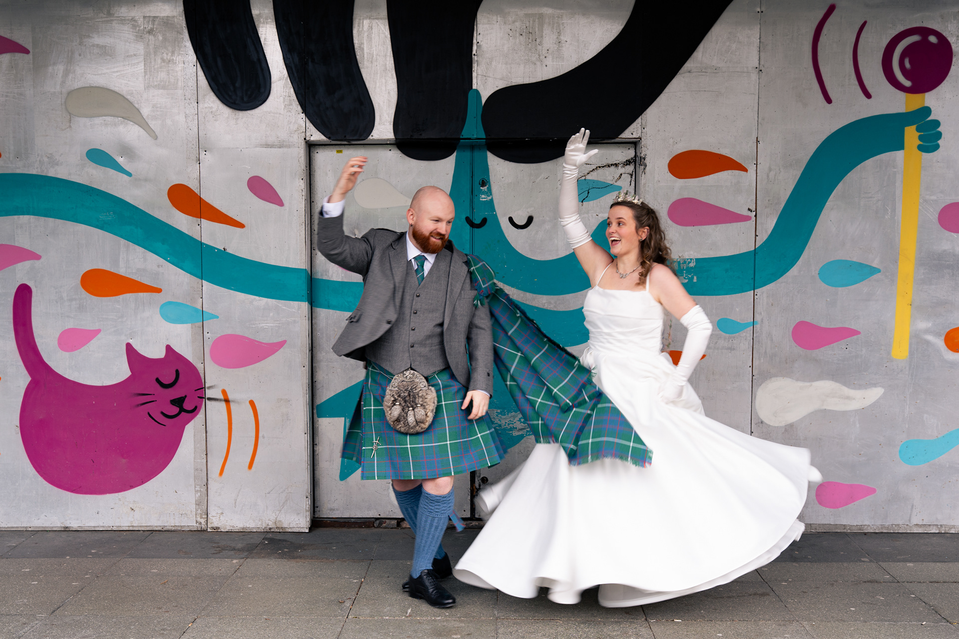 Bride in white and groom in kilt spin and laugh in front of graffiti in Aberdeen City centre during their laidback wedding.