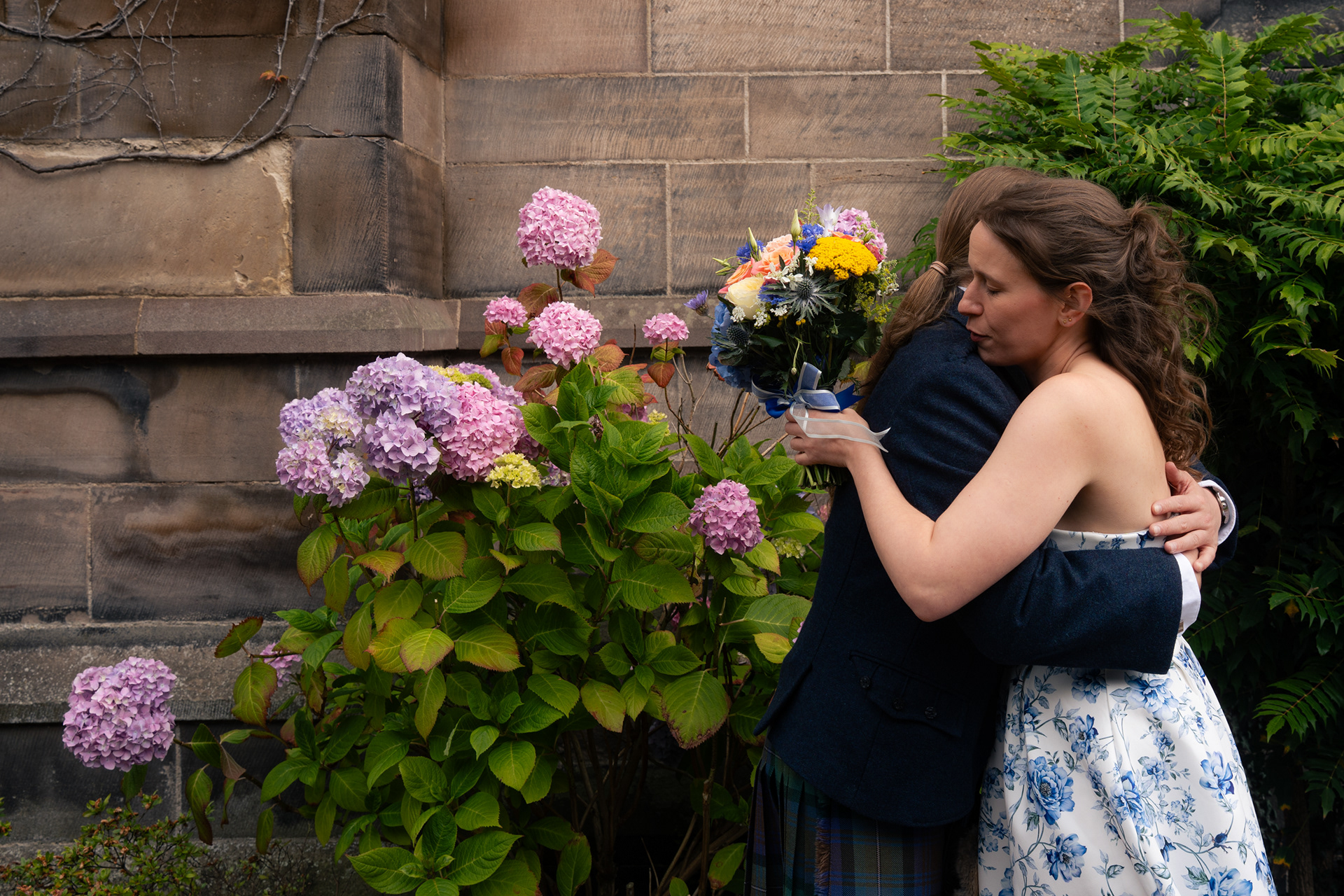 Aberdeen Photographer for Small Weddings: A bride in blue and white floral dress and a long-haired groom in navy blue hug each other tightly against a backdrop of hydrangeas. Her eyes are closed and his face isn't visible. The moment feels tender and protective.
