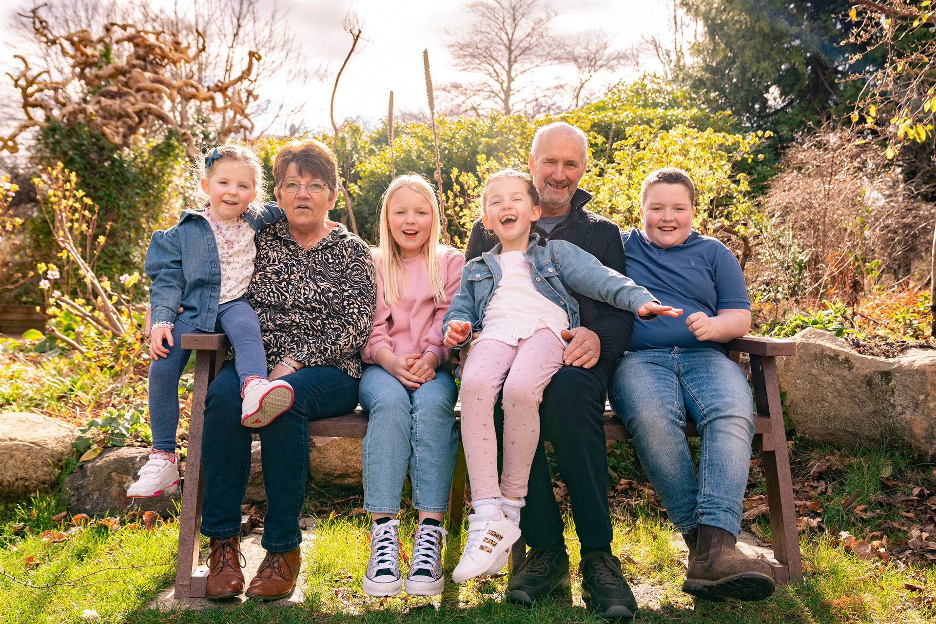 At an Aberdeen family photo shoot, two grandparents and four grandchildren in shades of pink and blue laugh together on a bench in a springtime park.