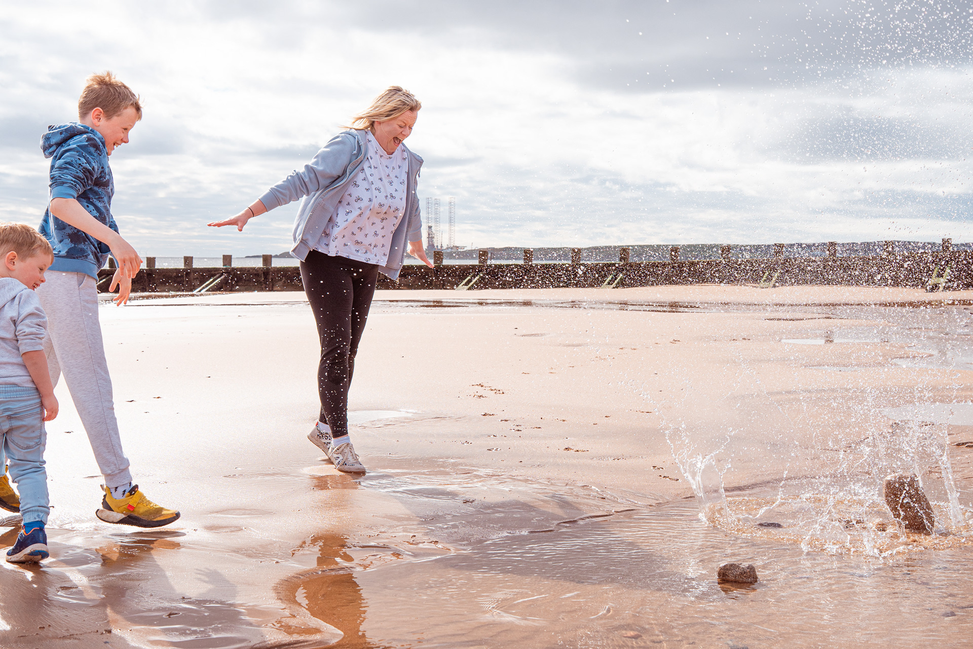 Aberdeen Family Photography: A mum in a floral top and dark leggings jumps back and shrieks as her two young sons, dressed in blue, throw a rock into the water and it splashes up around her. Everyone is laughing.
