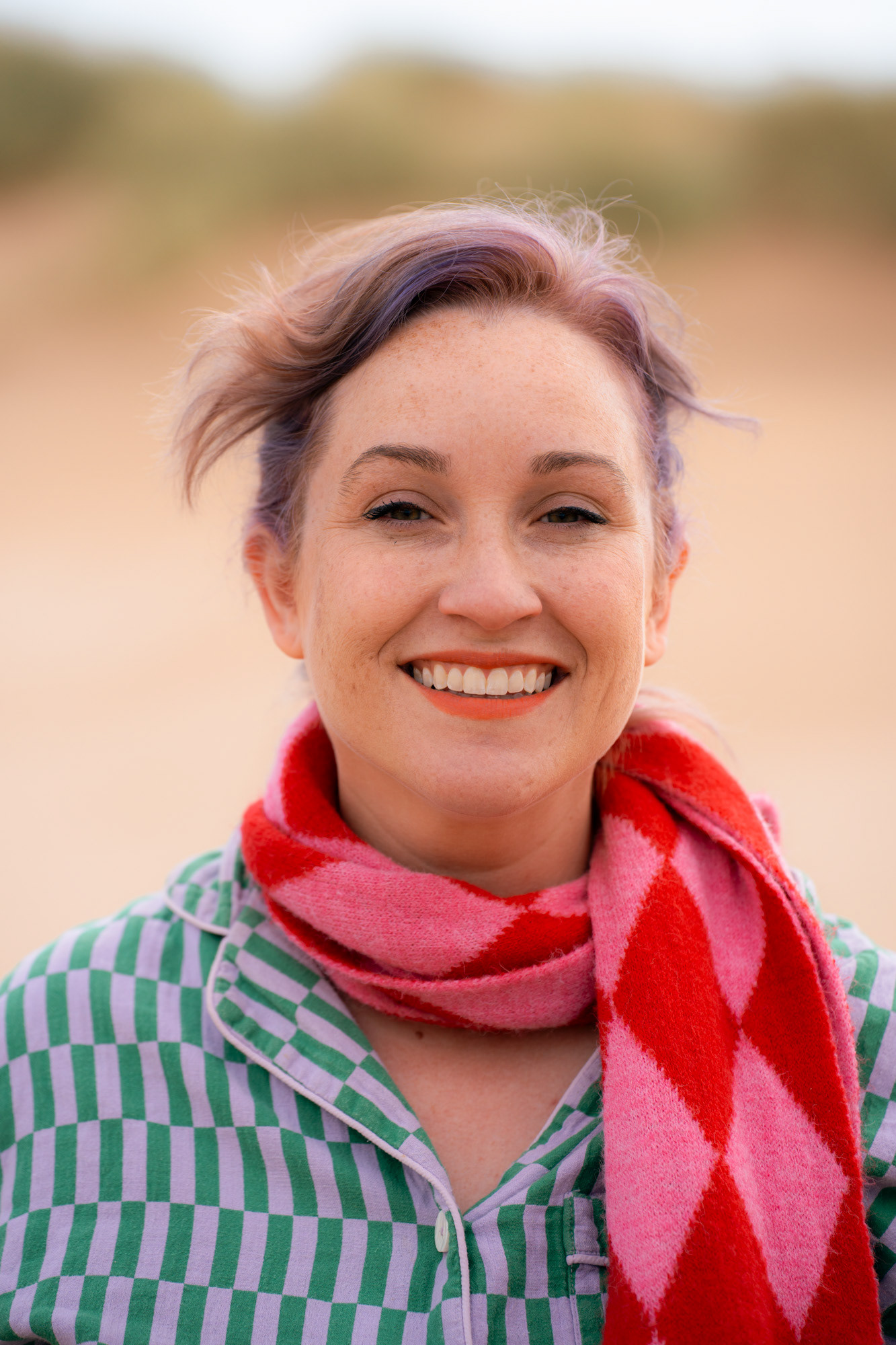 A white woman with ginger and lilac hair and freckles smiles directly at the camera.
