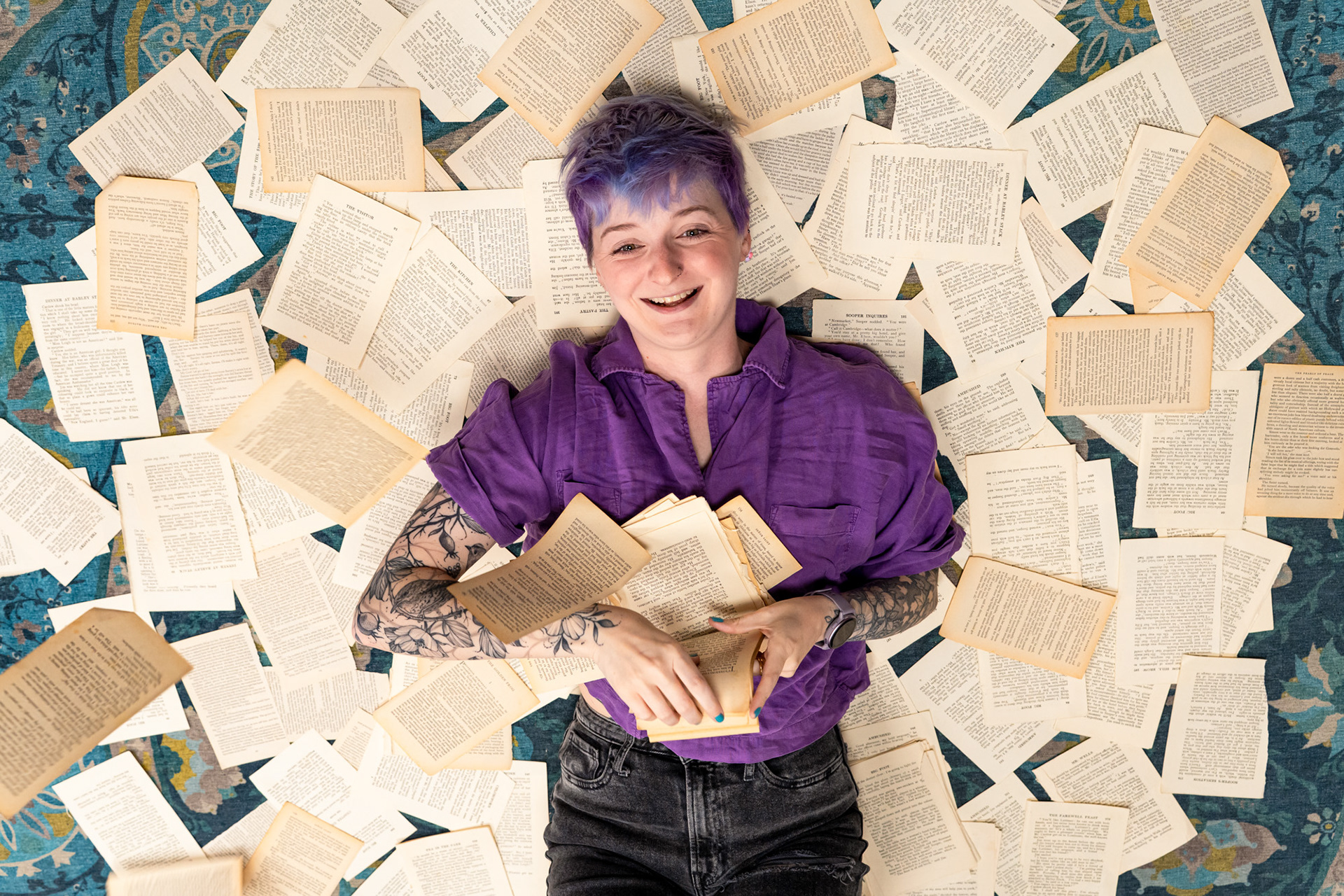 Bookshop owner, Christie, a white woman with short purple hair and amazing bird, cat and floral tattoos laughs as she lies on a floor covered in torn book pages and throws more in the air.