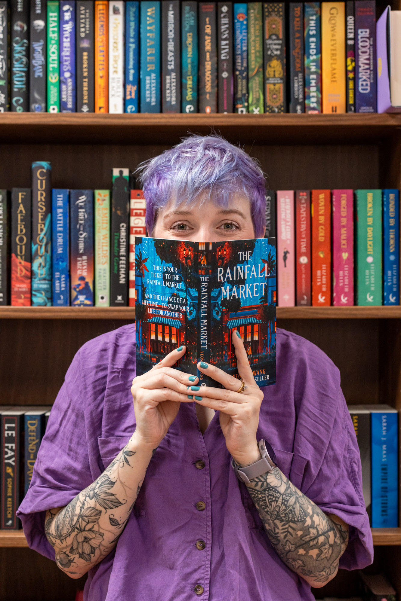 Bookshop owner, Christie, a white woman with short purple hair and amazing bird, cat and floral tattoos holds a book up over her face and peers above it. The backdrop is shelves of colourful books. Aberdeen branding photography session.