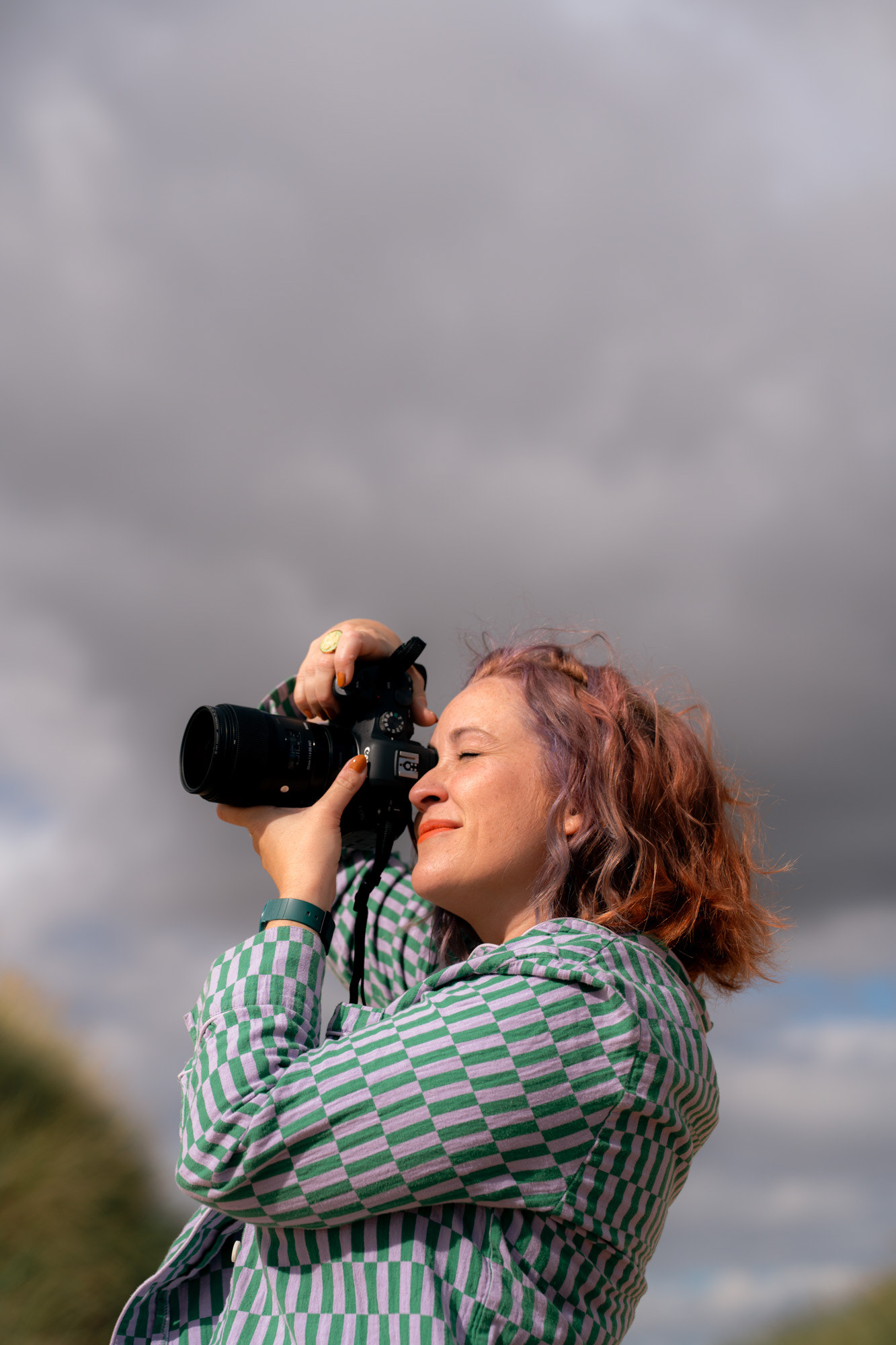 A white woman with ginger and lilac hair and freckles looks through the viewfinder of her camera, pointed off screen. The background is grey clouds, but she is lit by sunlight.