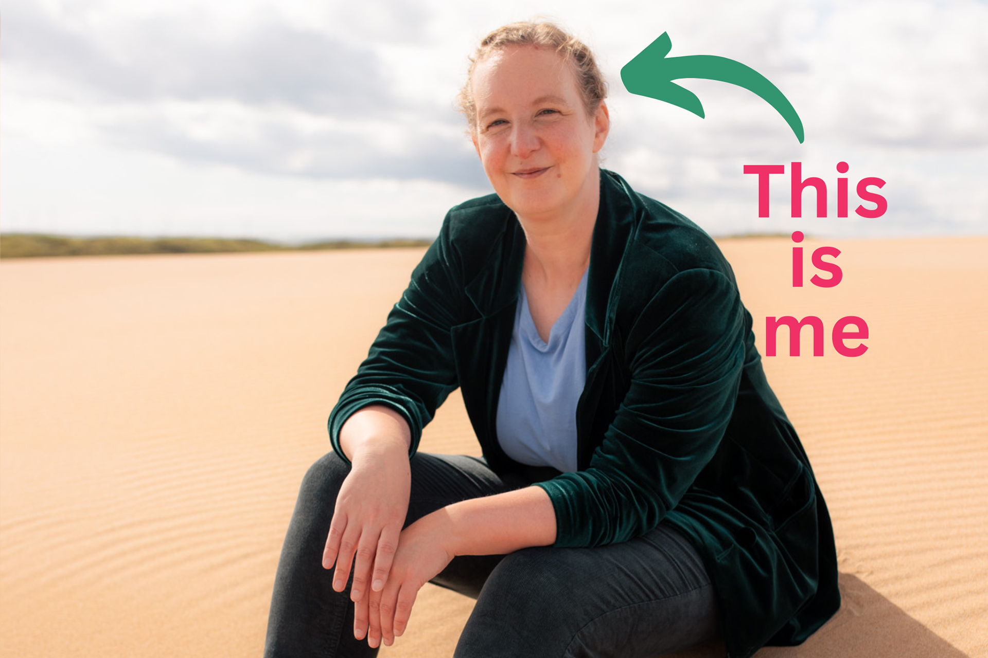 Aberdeen wedding photographer, Sarah from Windswept Stories, sits on a sand dune and smiles at the camera. She is white, middle-aged with curly hair tied back, and she wears a blue T-shirt with a green suit jacket, and is resting her wrists on her knees.