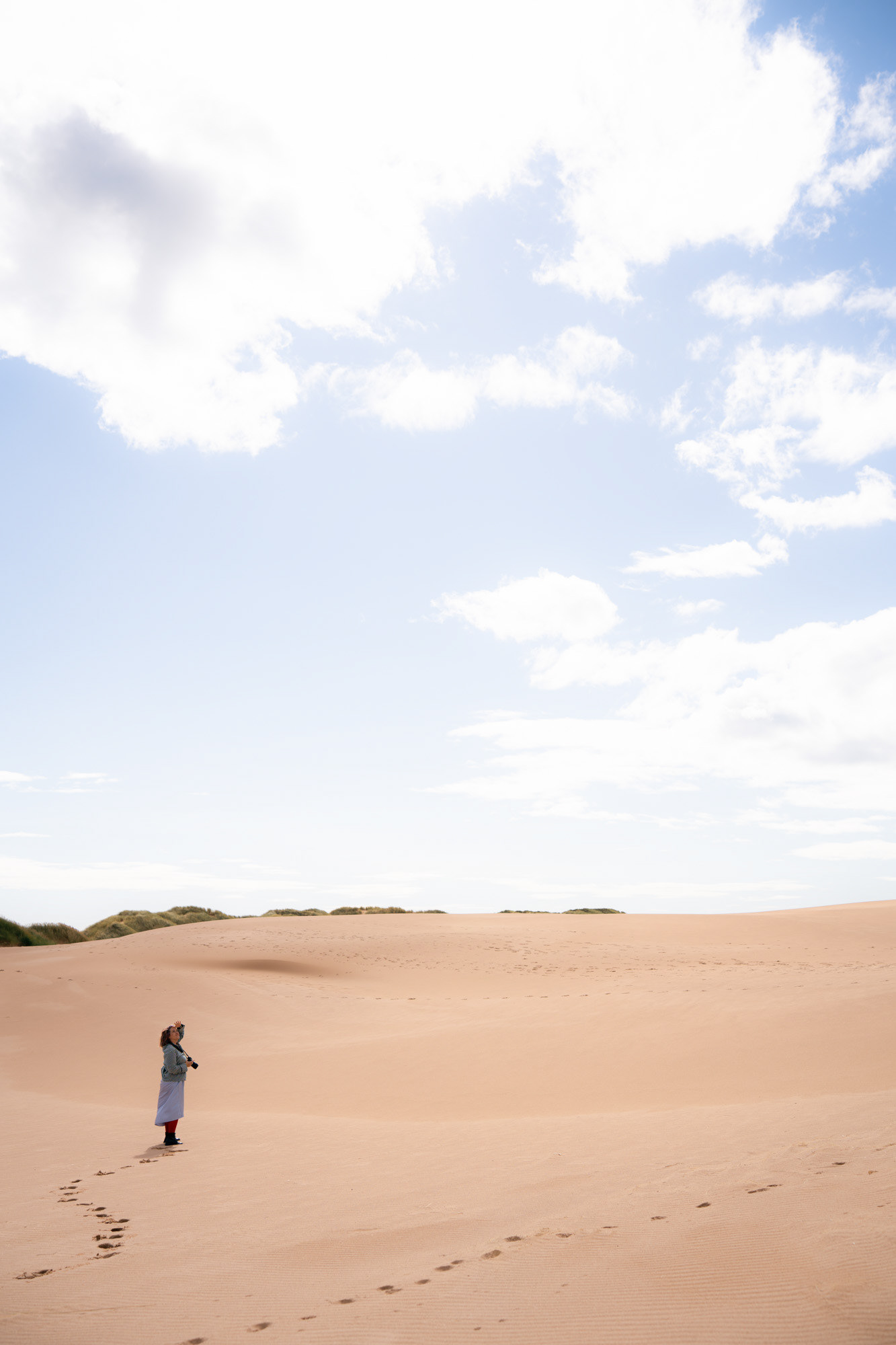 A woman in a lilac dress stands on a sand dune, her footprints tracking through the sand. She is tiny against the expanse of sand and the big blue sky above.