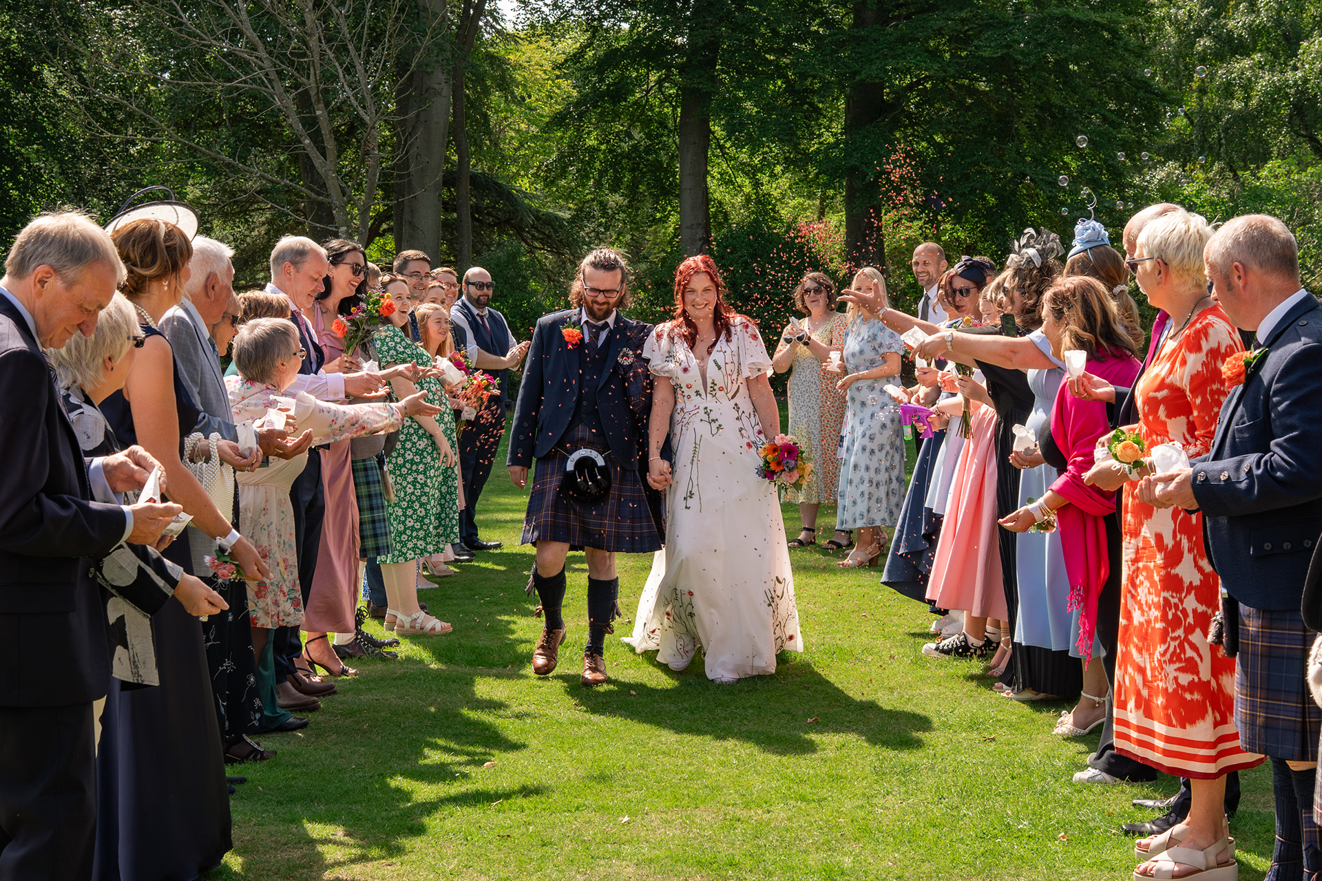 A newlywed couple - he in a kilt and she in a floral dress - walk between their guests at Hazlehead Park, being showered with pink confetti.