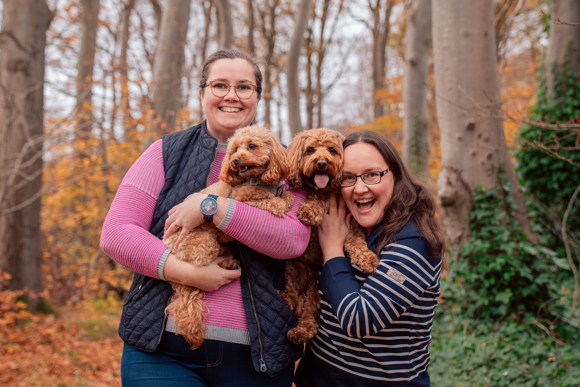 At their Aberdeen engagement photo shoot, two women in striped tops cuddle their beautiful dogs in an autumn forest.