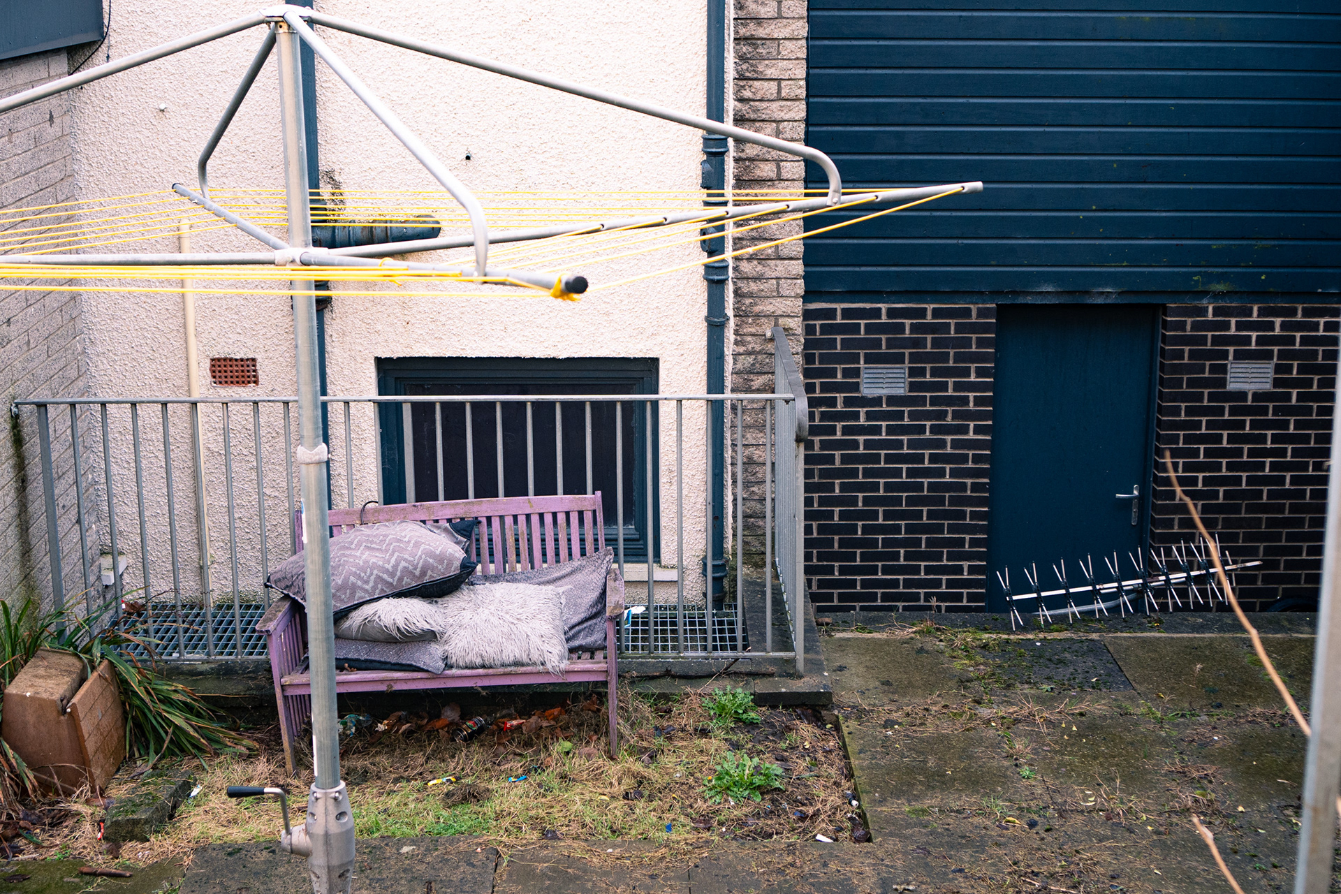 A lilac bench piled with rotten cushions sits in a patch of overgrown garden. A TV aerial lies nearby. A whirligig sits empty.