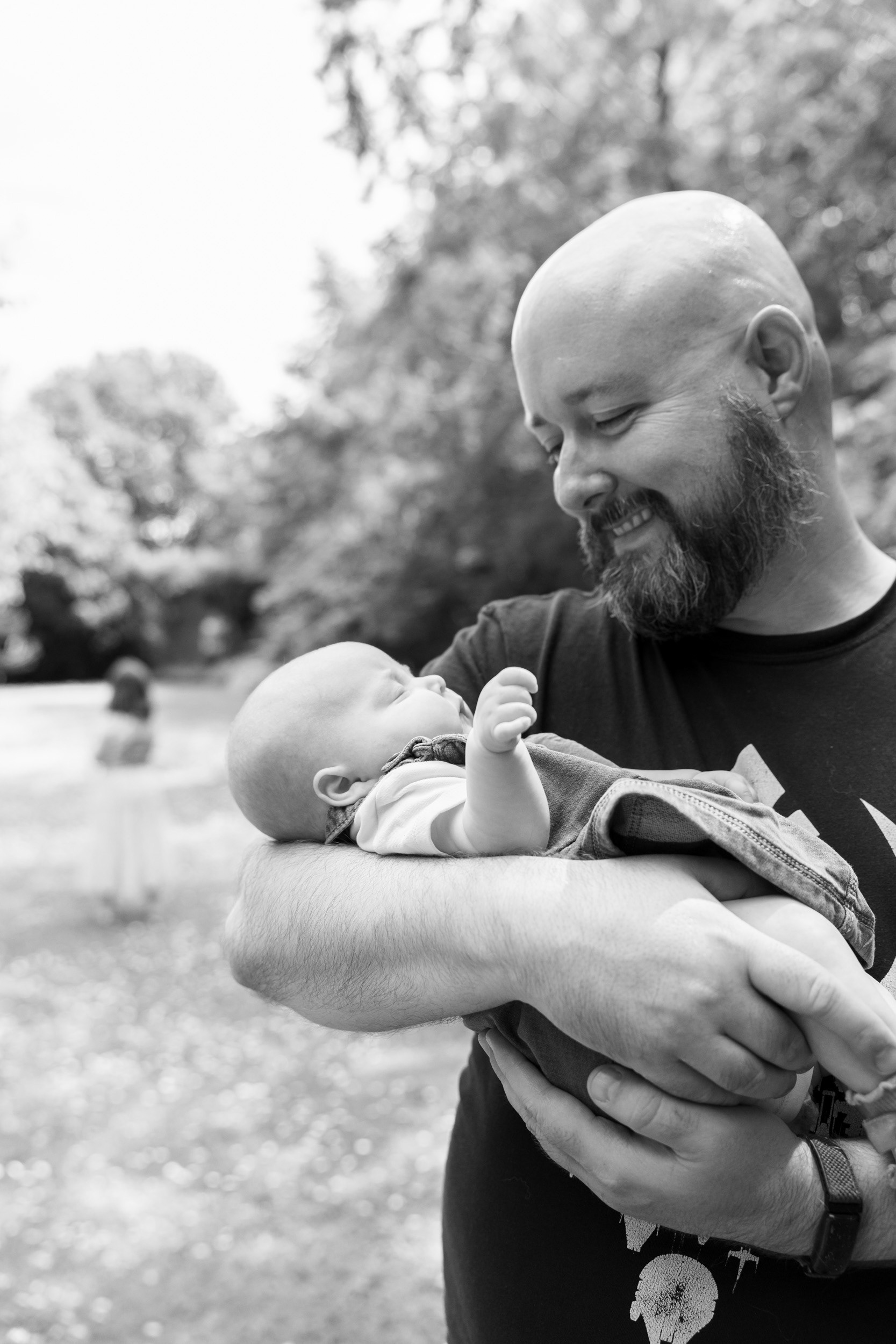 A black and white image of a dad with beard and shaved head cradling his newborn daughter and smiling, while his 7 year old daughter dances off in the background.