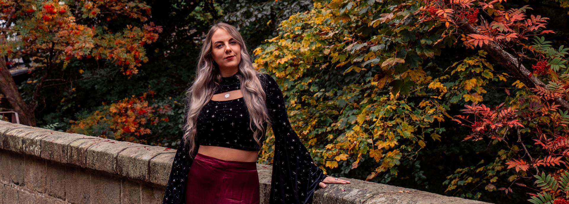 Taken during a gothic Aberdeen empowerment photo shoot, a white woman with long blonde hair, wearing a black starry top, looks directly at the camera. The background is red and yellow autumn leaves still on the trees.