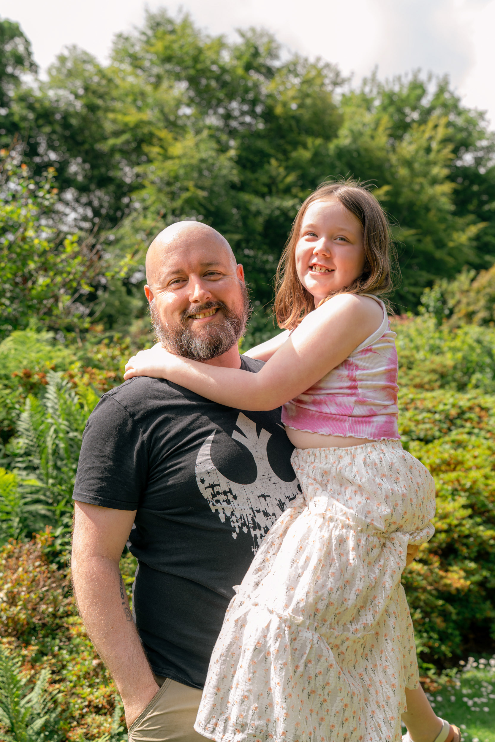 Alternative Aberdeen family photography: A dad stands lifting his beaming 7 year old daughter in a sunny garden.
