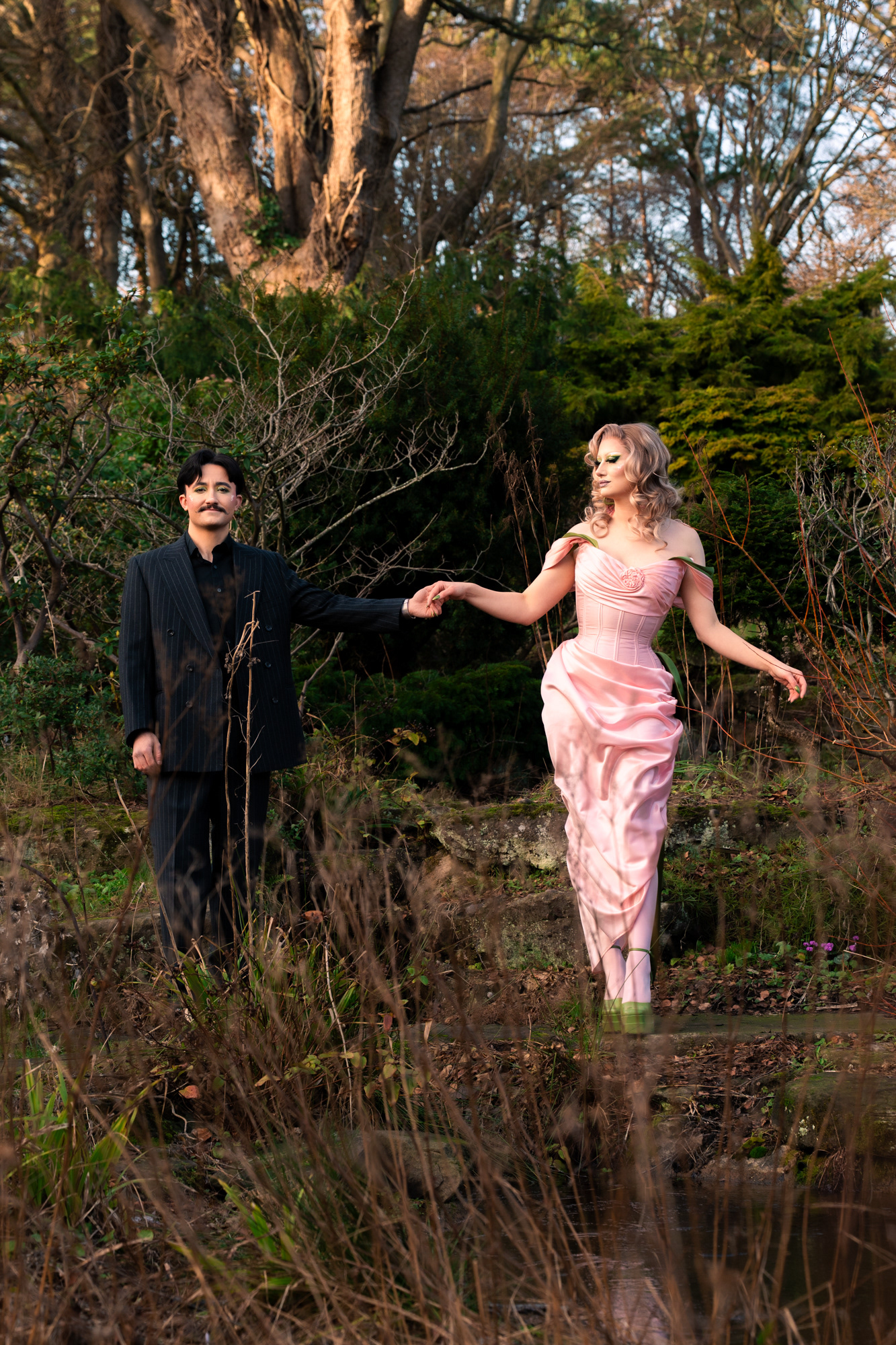 A couple in drag pose together during their Aberdeen couple's photo session. They/them is wearing a pinstripe suit and has short, dark hair and a moustache. She/her is in a glamorous pink and green dress and high green heels with dramatic makeup.  They are surrounded by greenery, holding hands; she is looking at them and they are looking at the camera.