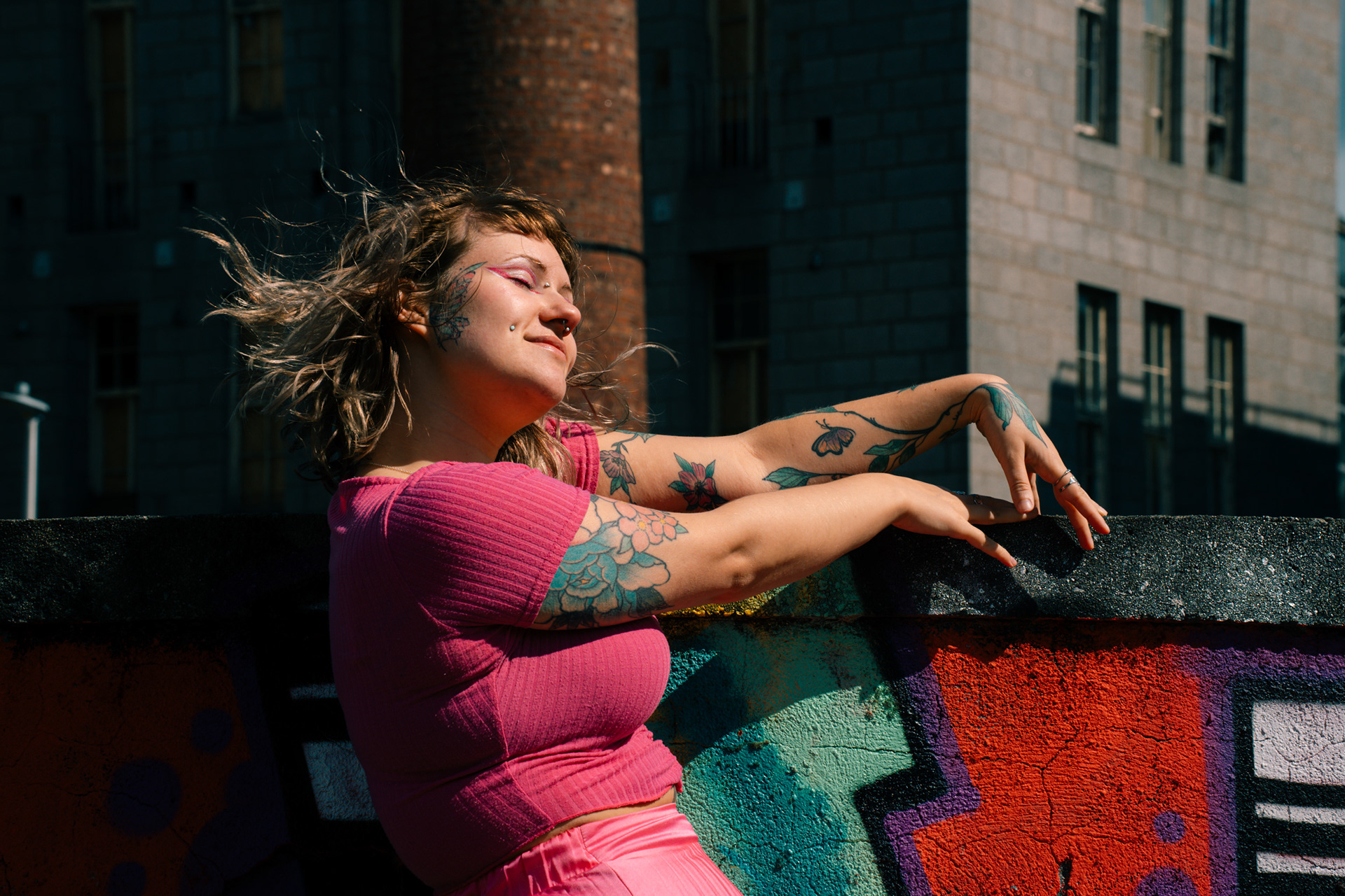 A young woman's short hair blows out behind her as she stands against a graffitied wall, head turned towards the sun. She is wearing a pink top and has tattoos and bold, colourful make up including a butterfly across her cheek.