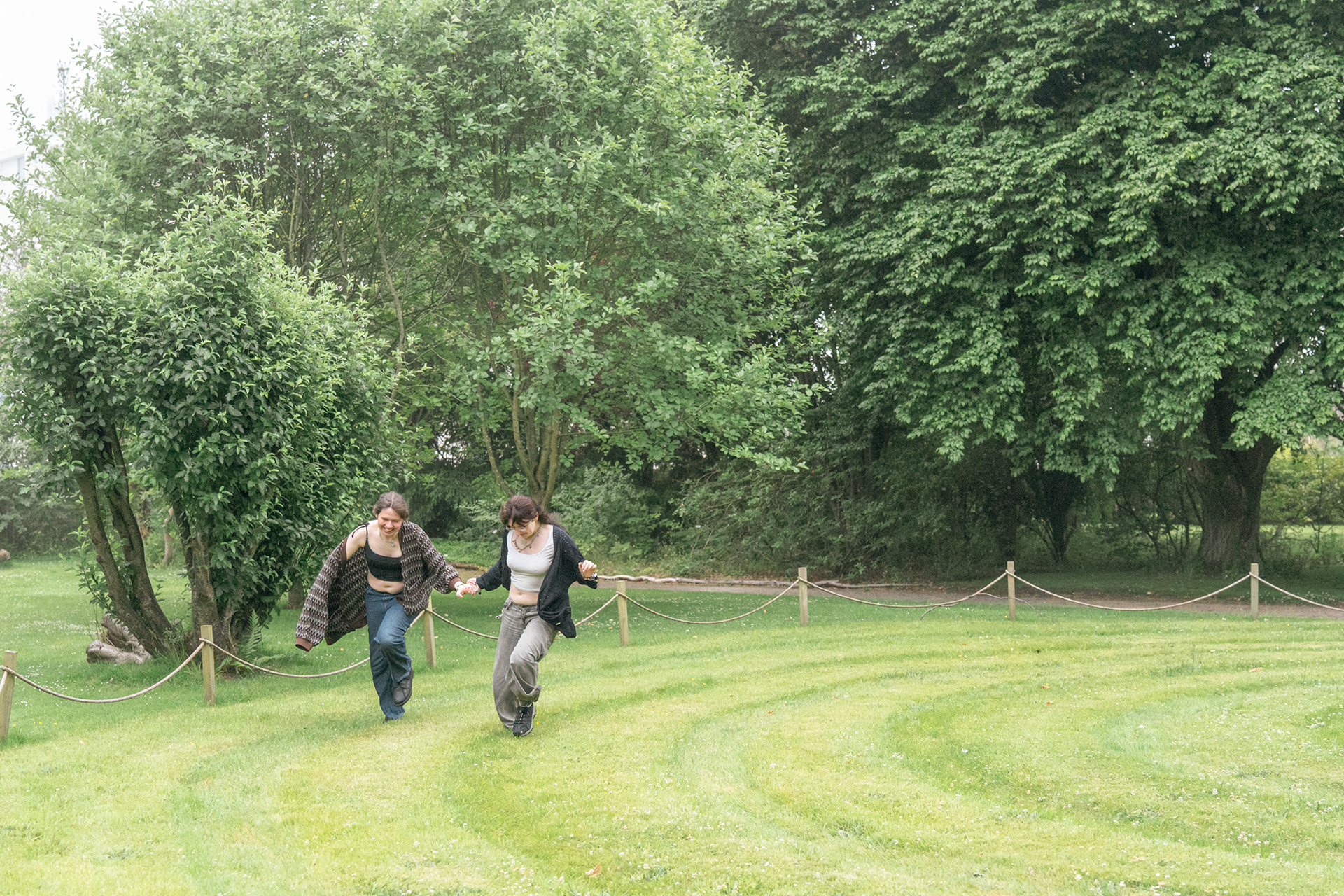 A young couple in jeans and long cardigans hold hands and laugh as they run around a spiral mown into a lawn in an Aberdeen garden. Their cardigans are flying out behind them. Photo taken by LGBTQ+ Aberdeen wedding photographer, Sarah.