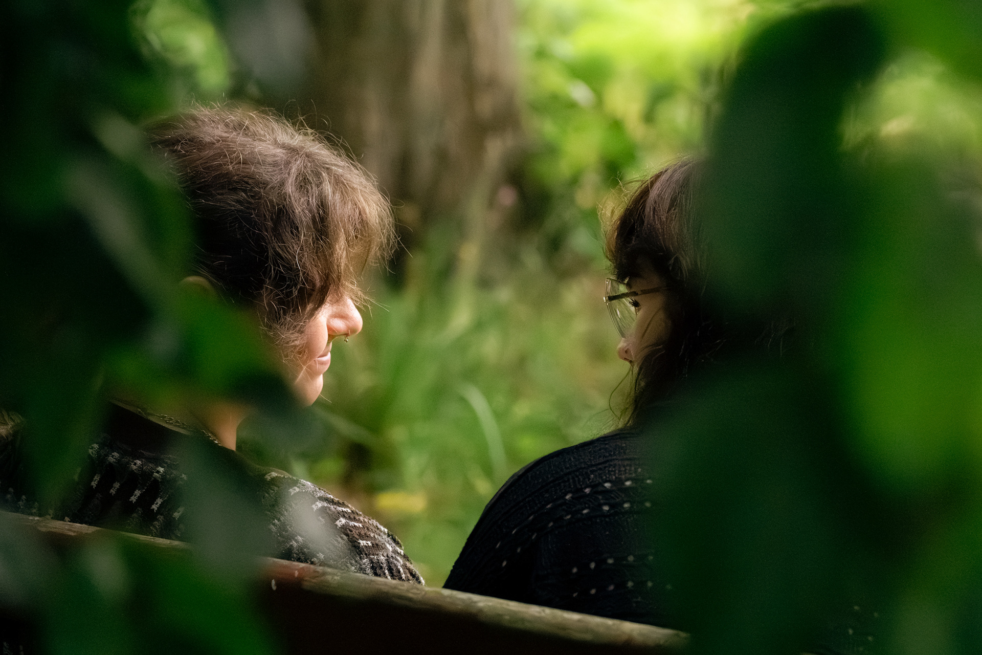 Two wives with long brown hair, face each other, apparently mid-conversation, during their Aberdeen couple's photo shoot. They are photographed from behind, with leaves framing their faces in the foreground.