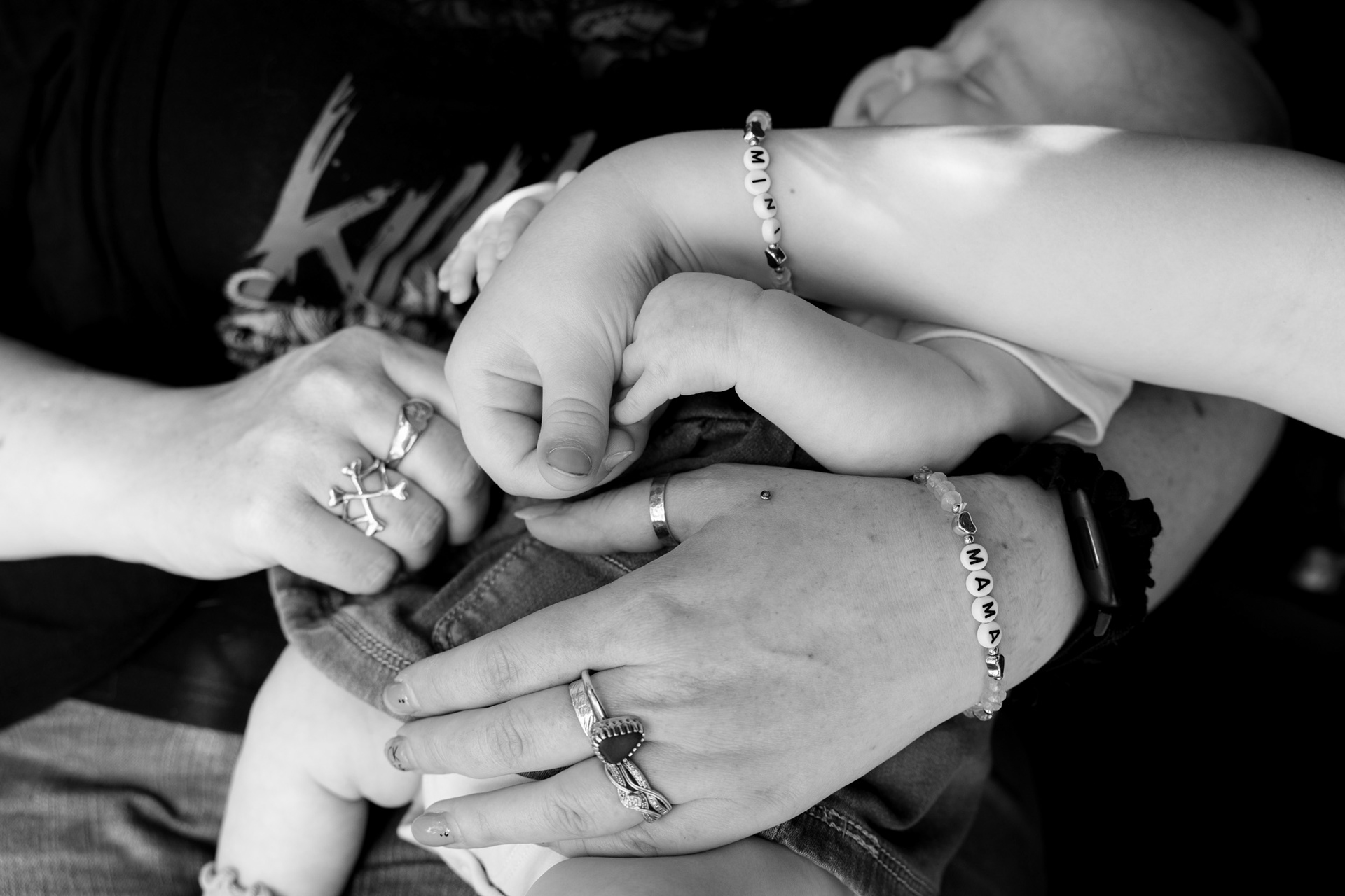 Aberdeen Family Photography: A black and white image of a sleeping baby with mum and big sister's hands wrapped around her. Mum wears a beaded bracelet reading "MAMA" and big sister has a matching one reading "MINI".