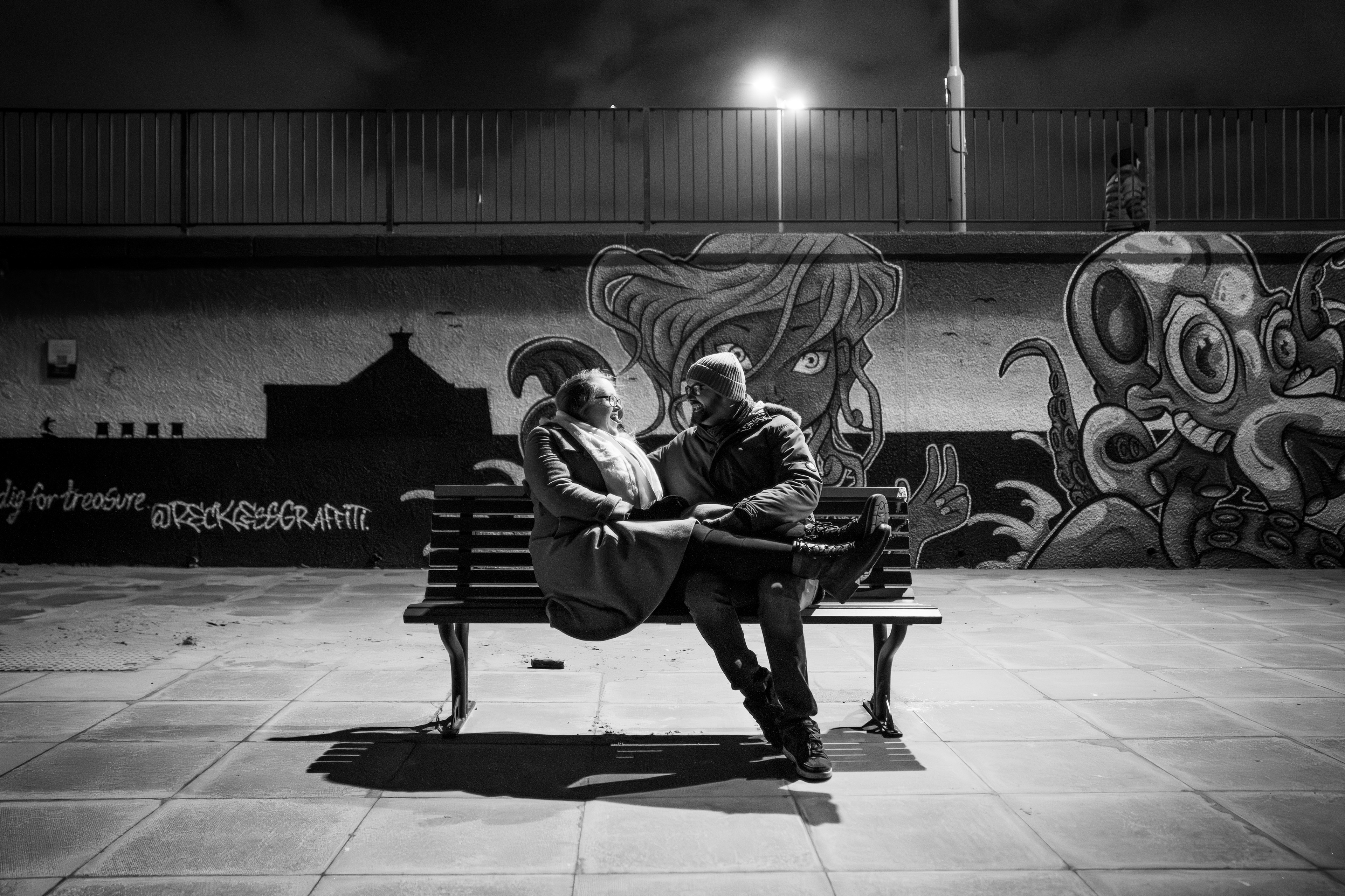 Black and white image: On an nighttime photo shoot at Aberdeen beach, a cis-het couple sit on a bench under lamplight, smiling at each other. They are dressed in winter coats and the man wears a woolly hat. The background shows alien street art.
