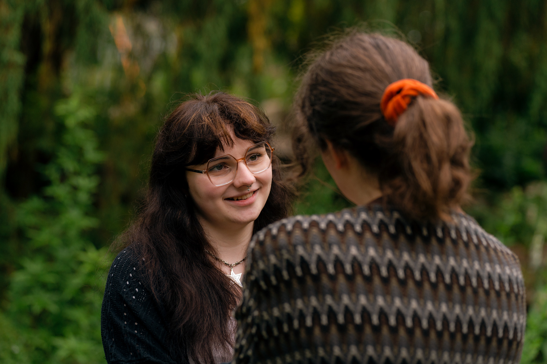 A non-binary person with long dark hair and glasses gazes at their wife, who we see from behind.