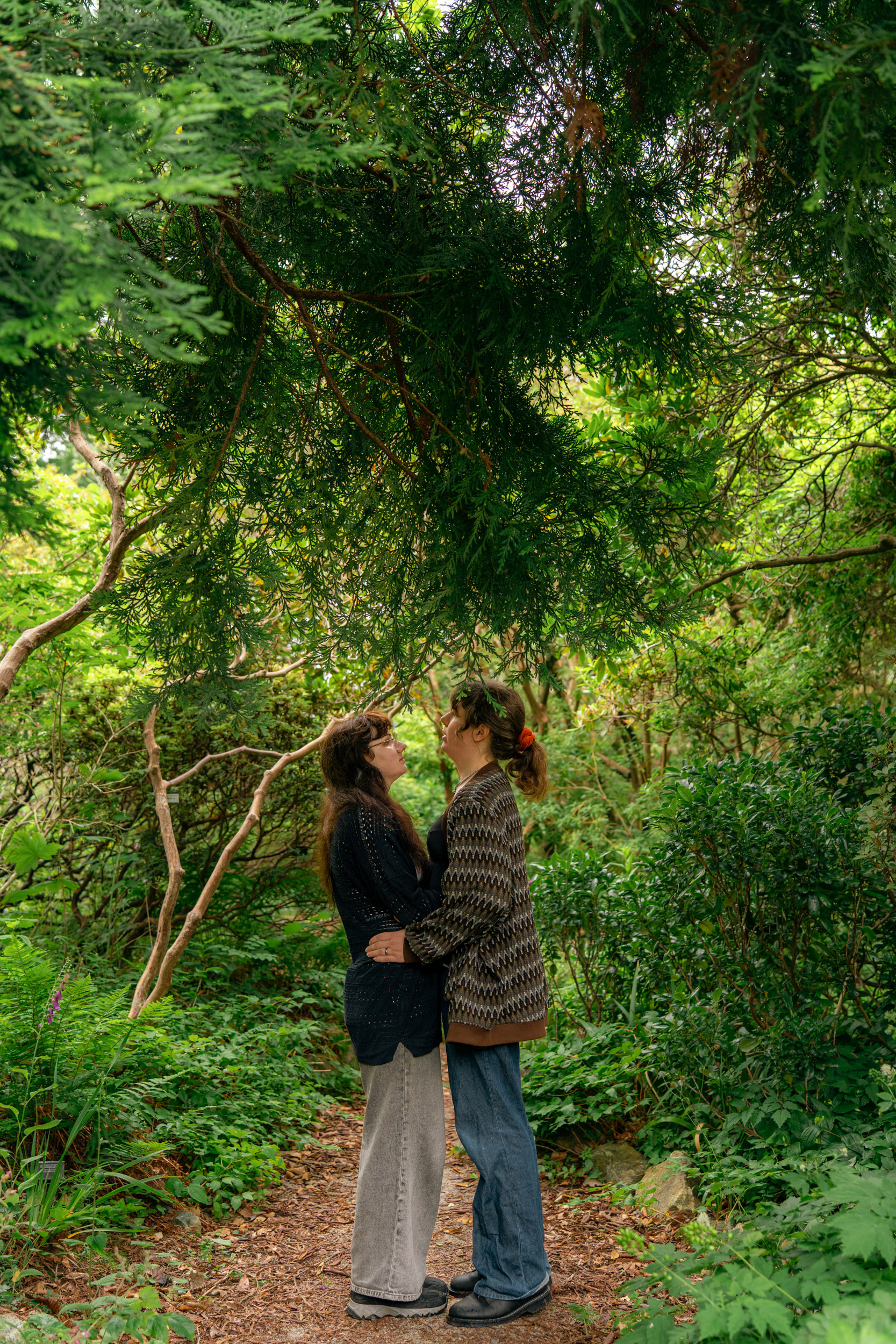 In an Aberdeen garden, trees and ferns frame a married couple (she/her and they/them) as they stand with their arms around each other, face to face.