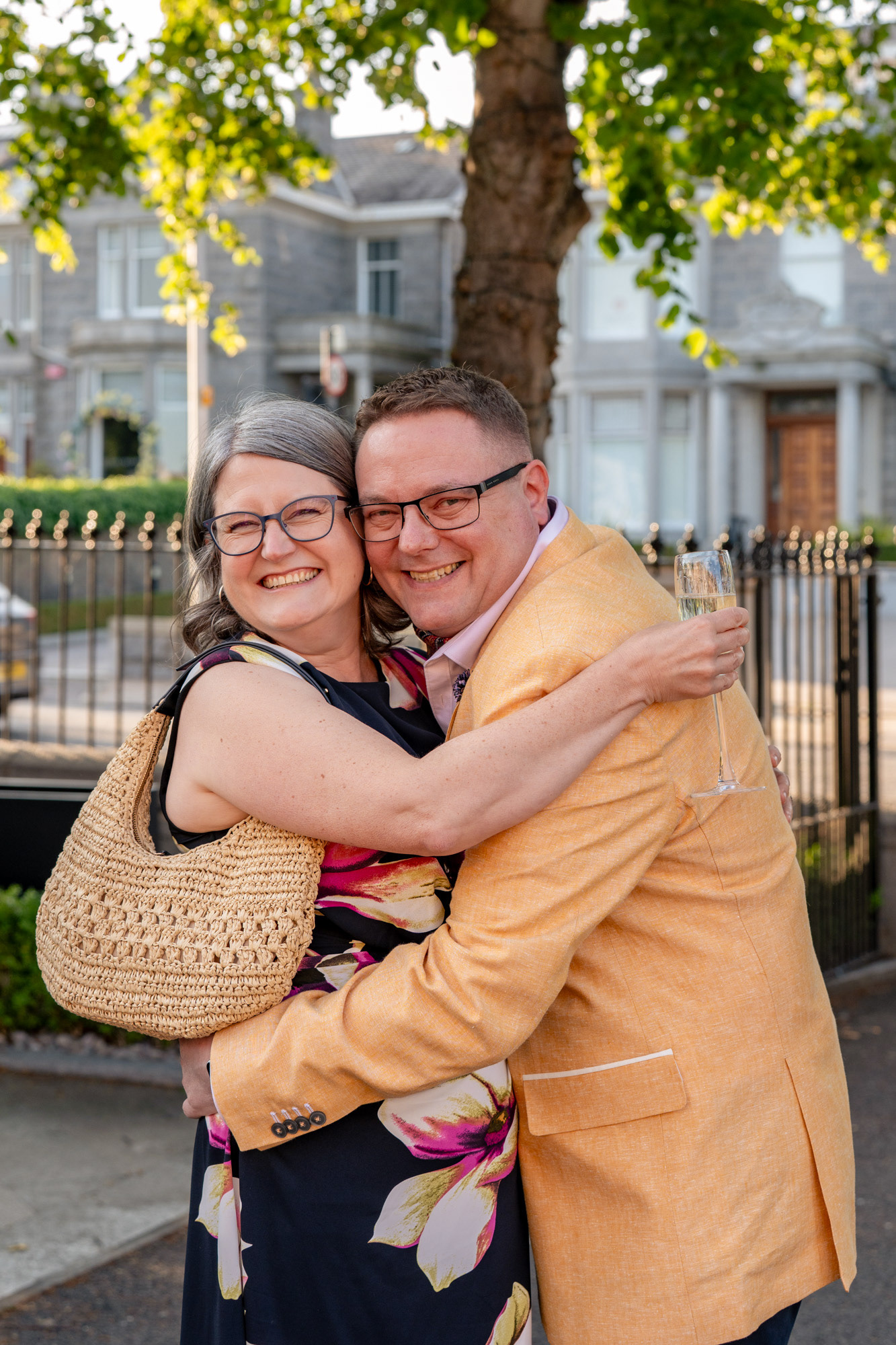 A man in a yellow jacket hugs a woman in a black floral dress and they both laugh and smile directly at the camera outside Aberdeen's Chester Hotel.