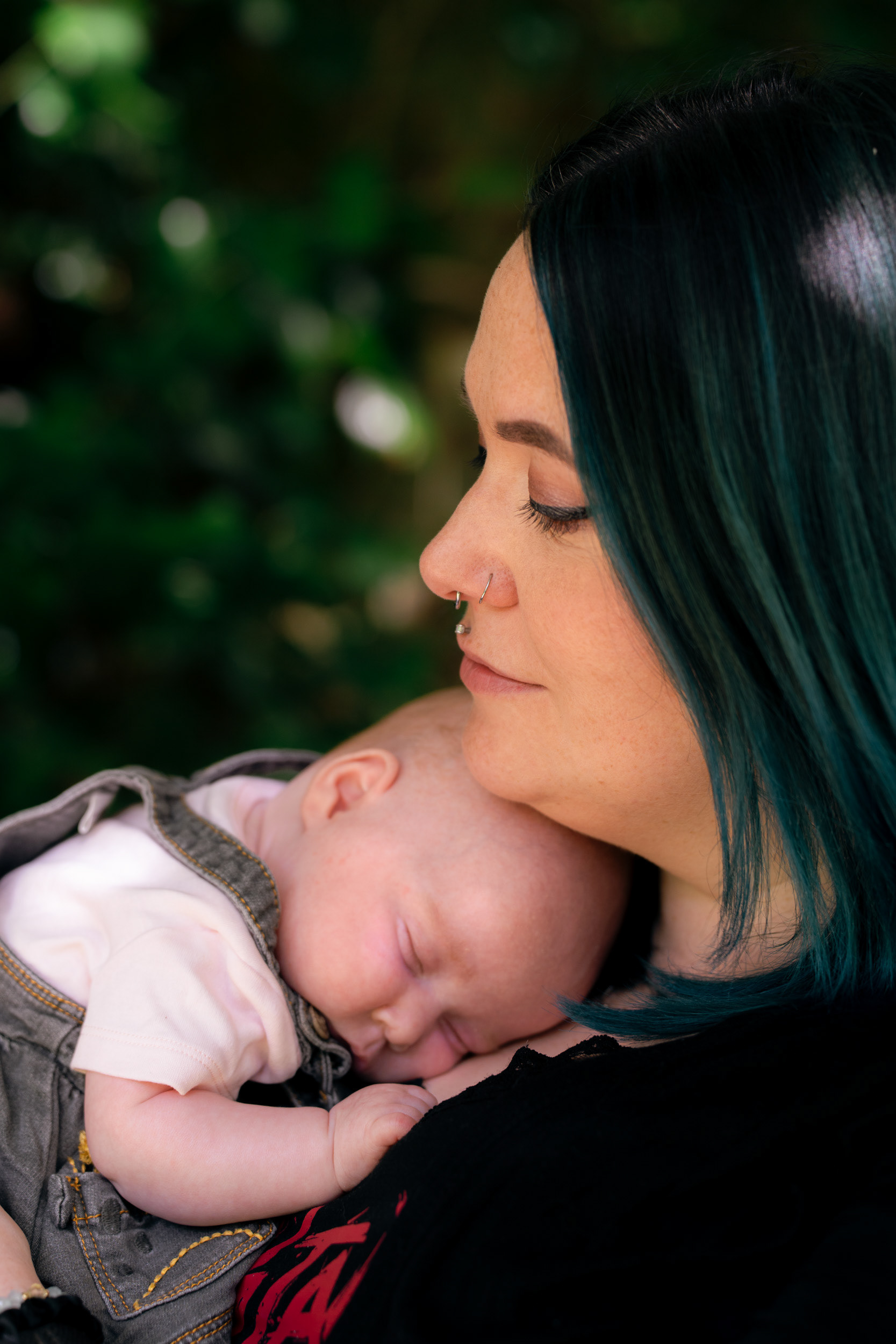 A mother with black and green hair and facial piercings, looks down at her baby daughter, curled up sleeping under her chin, during an Aberdeen newborn photo shoot.