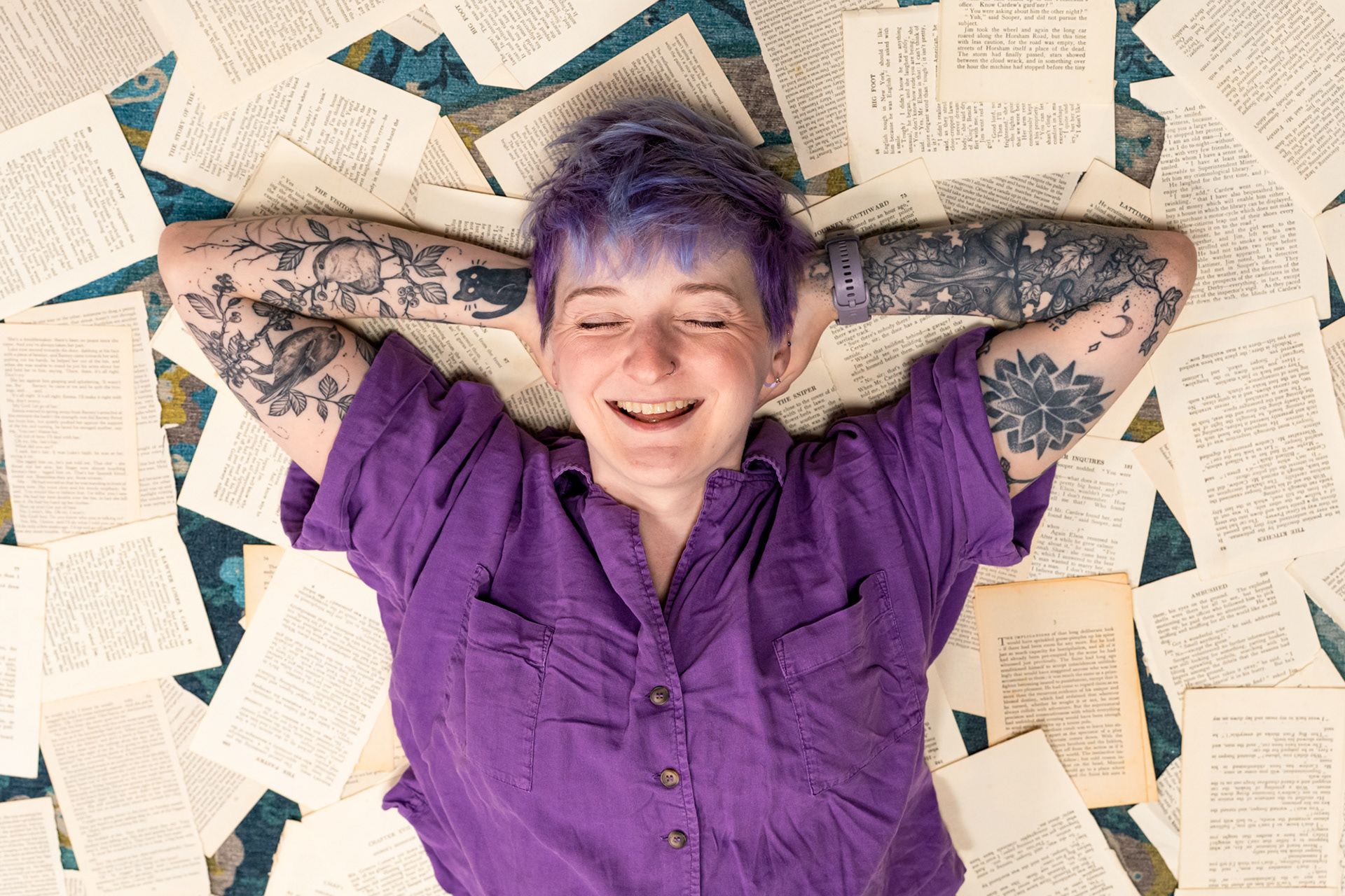Bookshop owner, Christie, a white woman with short purple hair and amazing bird, cat and floral tattoos lies on a floor covered in torn book pages, eyes closed, hands behind her head, laughing.