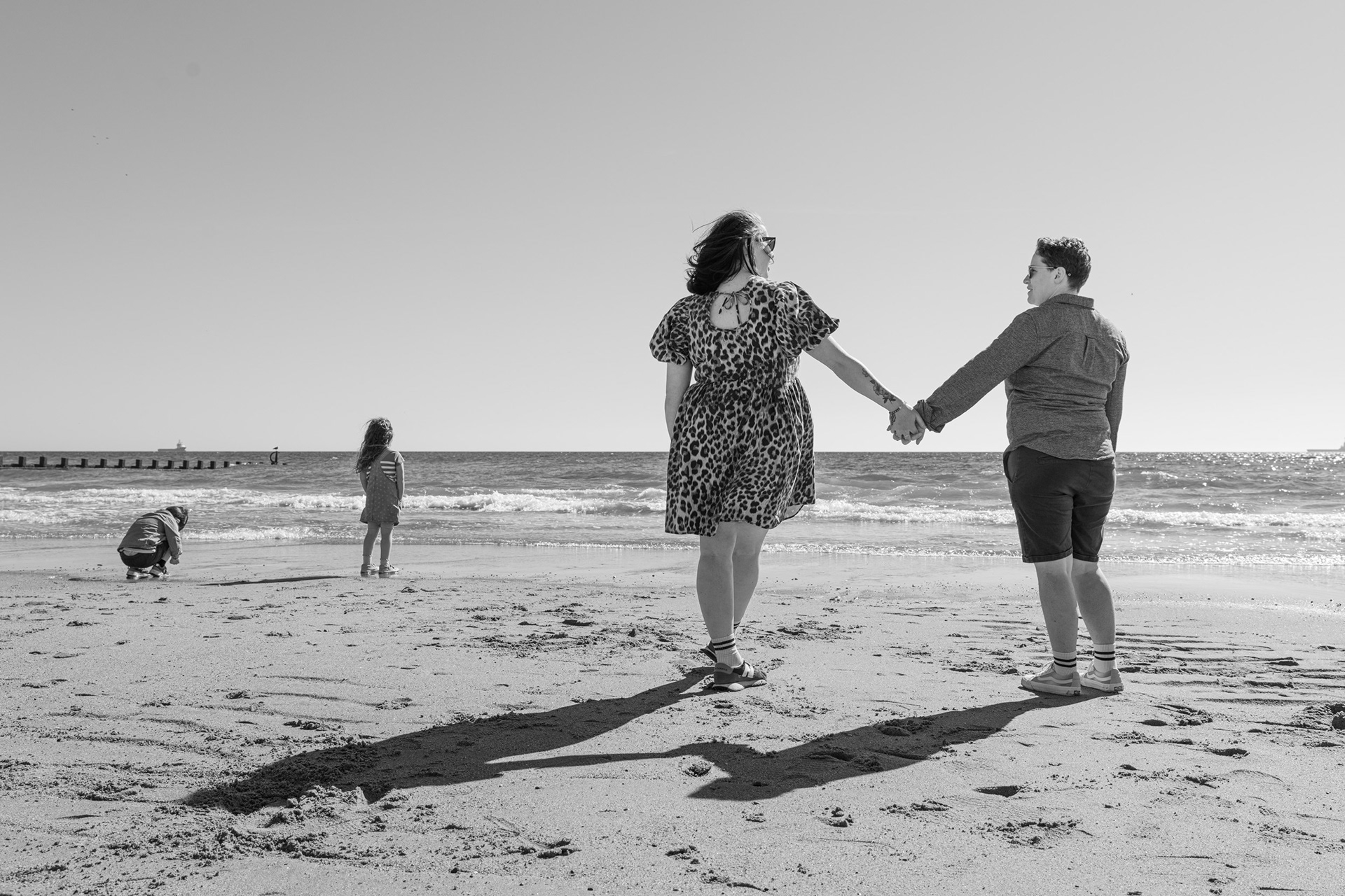 Aberdeen family photography for queer (and hetero) love. Two mums stand, hand in hand, looking at each other while their children play on the beach in the background.
