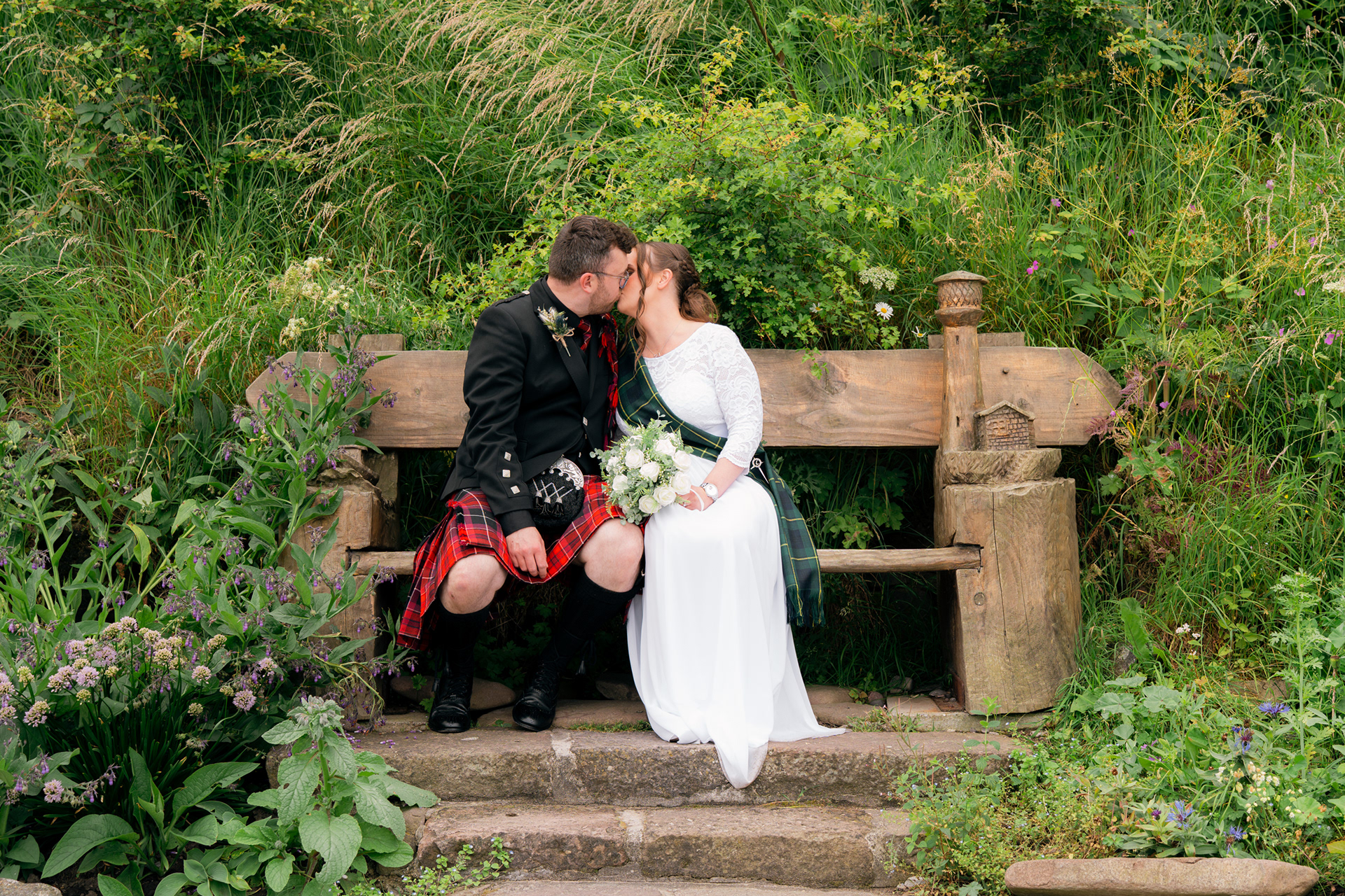Alternative Aberdeenshire wedding photography: Bride and kilted groom sit on an ornate wooden bench surrounded by wildflowers, kissing.