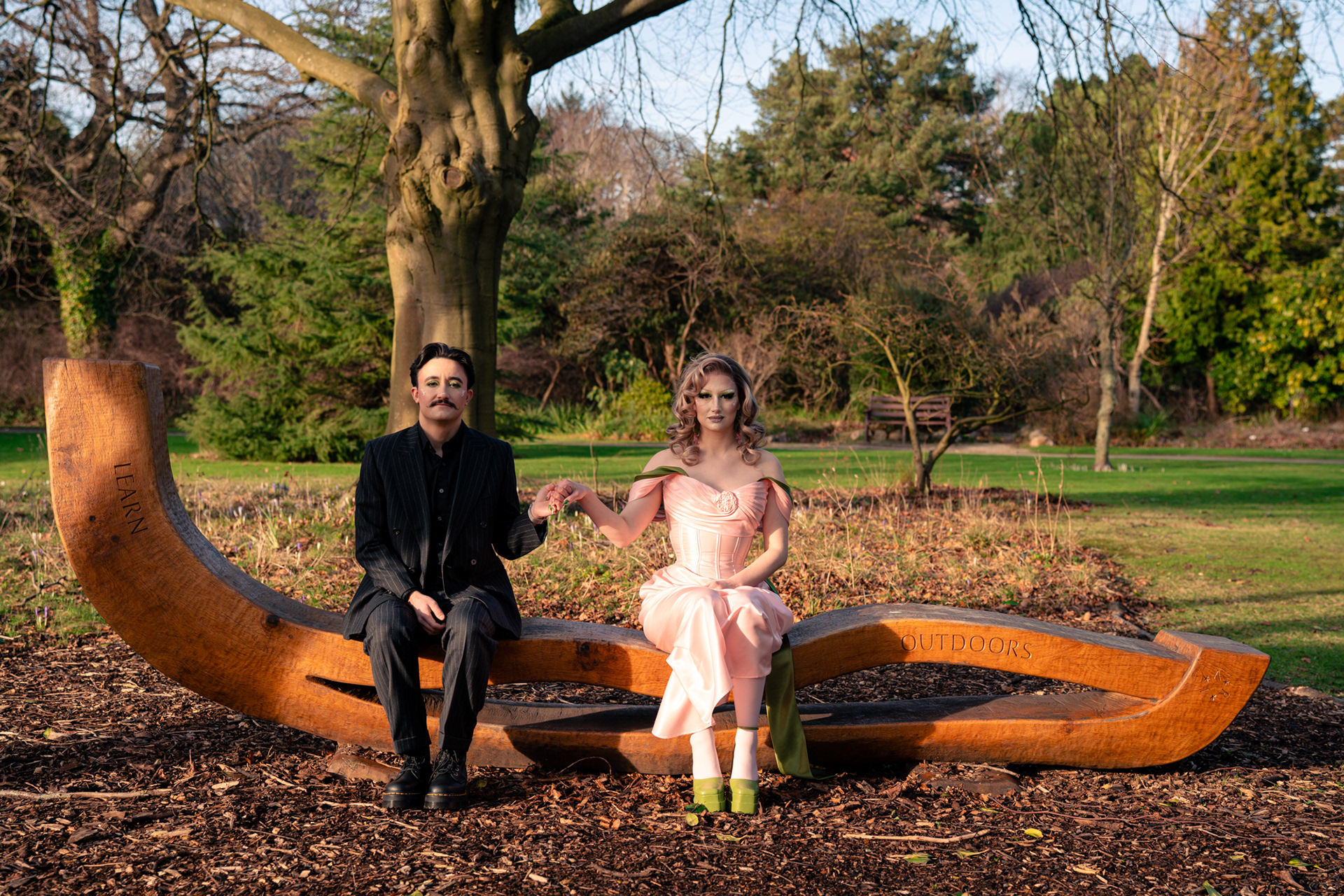 A couple in drag pose together during their Aberdeen couple's photo session. They/them is wearing a pinstripe suit and has short, dark hair and a moustache. She/her is in a glamorous pink and green dress and high green heels with dramatic makeup. They are sitting together on an unusually shaped bench, holding hands and looking straight at the camera.