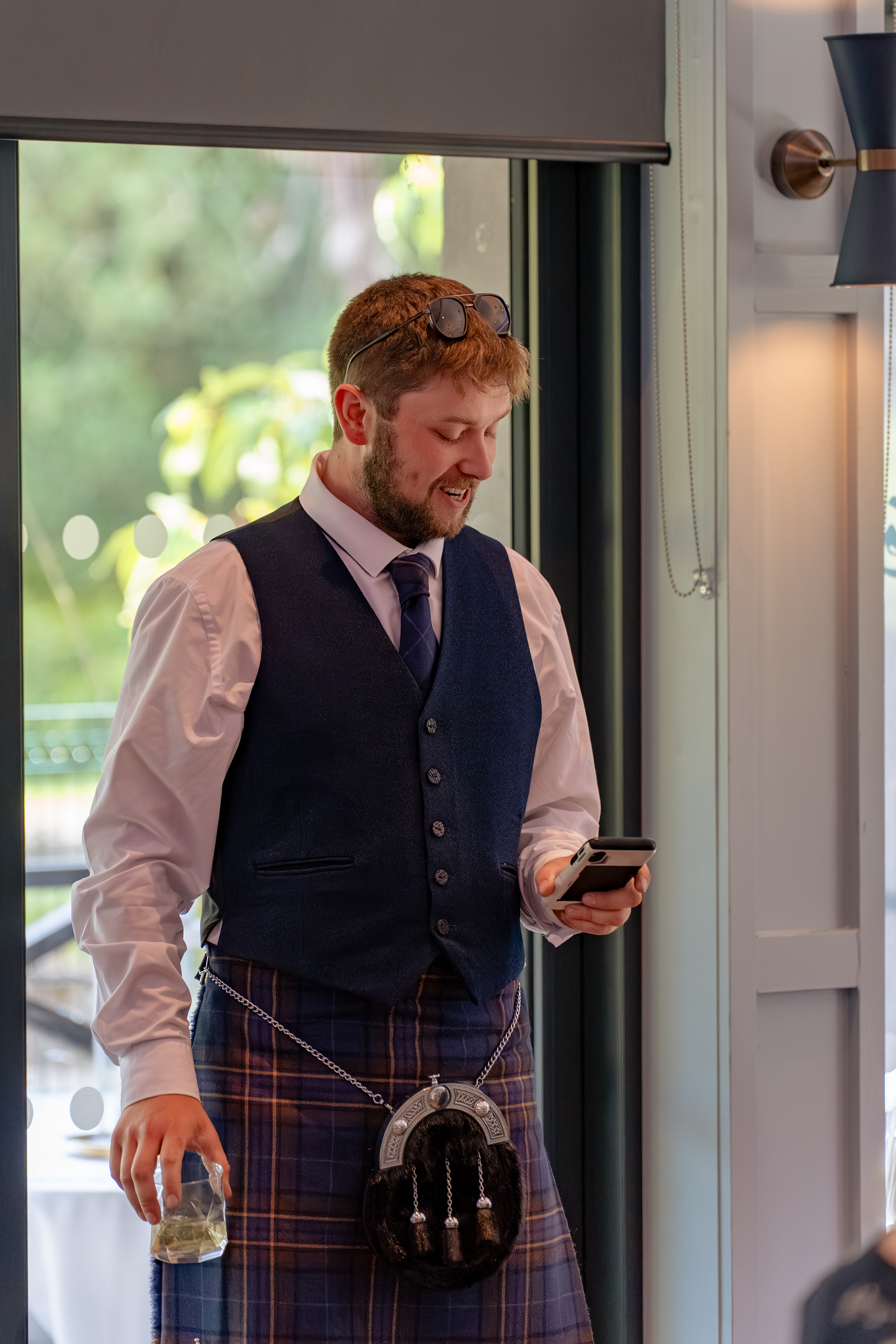 A young man in a kilt, with a short beard and sunglasses propped on his head, reads his wedding speech from his phone.