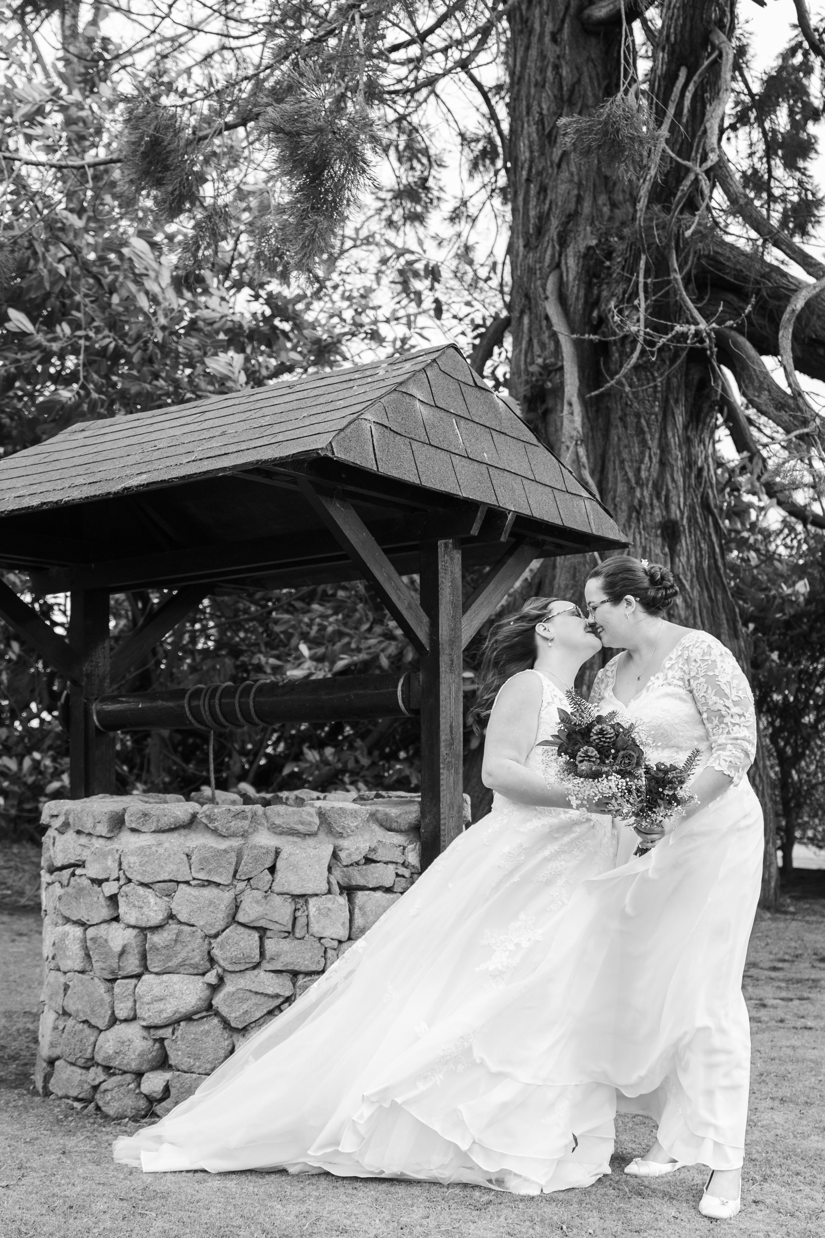 Black and white photo: Two brides in long white dresses, holding bouquets, kiss in front of the Marcliffe wishing well during their Aberdeen wedding.
