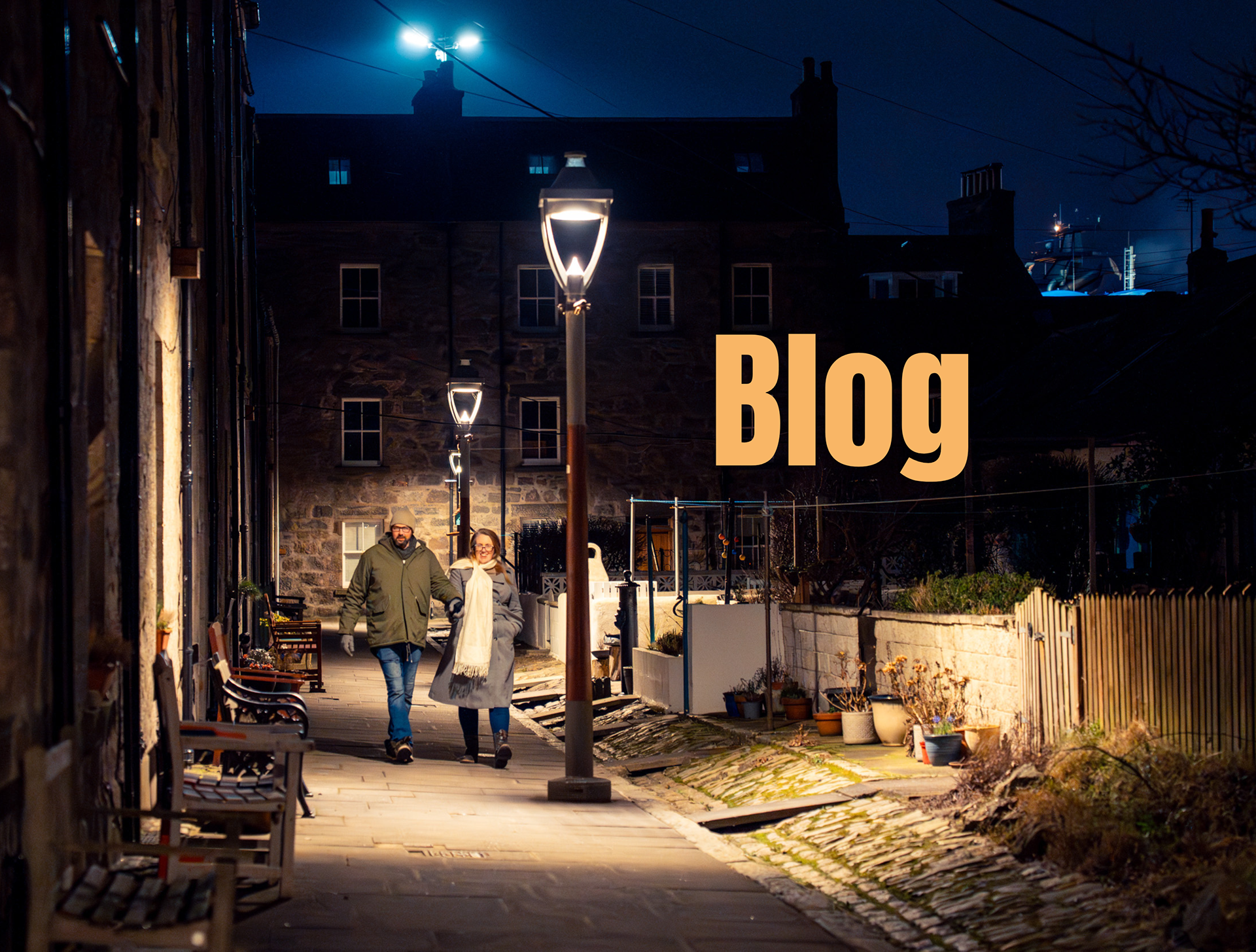 A couple in winter coats and woollens stroll through Fittie after dark. They are lit by the glow of an old fashioned street lamp. The overlaid text reads "Blog".